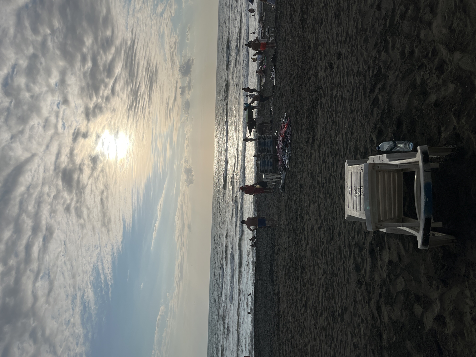 The image depicts a beach scene in Ureki, Georgia, during what appears to be late afternoon or early evening. The sky is filled with a mix of clouds, with the sun partially obscured and casting a bright glow. The sea is visible with moderate waves, and the beach is covered in dark sand.
In the foreground, a white plastic beach chair is positioned facing the sea. A clear plastic water bottle is attached to its side. The beach is populated with several individuals. In the middle ground, people are walking along the shoreline, some are wading in the shallow water, and others are engaged in activities like carrying surfboards. Beach umbrellas and towels are scattered on the sand.
Further back, more people are visible, some lounging on the sand, others in conversation. The overall atmosphere suggests a relaxed and recreational environment. The dark sand is a distinctive feature of the Ureki beaches, known for their therapeutic magnetic sand.