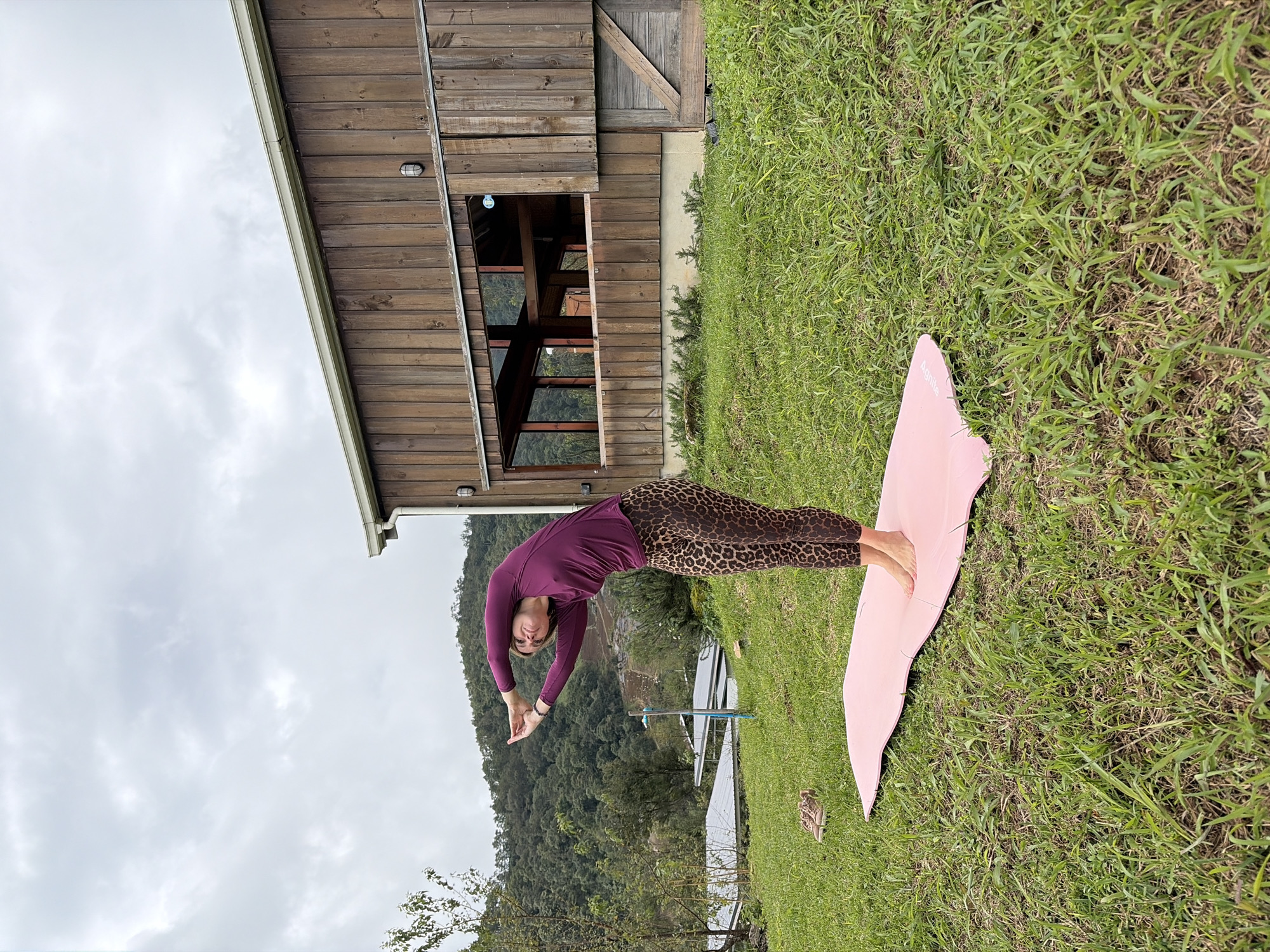 A woman with blonde hair is performing a yoga pose outdoors in Ban Khun Wang, Thailand, on a cloudy day. She is wearing a long-sleeved purple shirt and leopard-print leggings. She stands on a pink yoga mat placed on a grassy hill. Her arms are raised above her head, interlaced, and her body is arched backward. Behind her is a wooden building with a large window, and in the distance, there are lush green hills covered in trees. A pair of tan sneakers are visible on the grass to the left of the yoga mat.