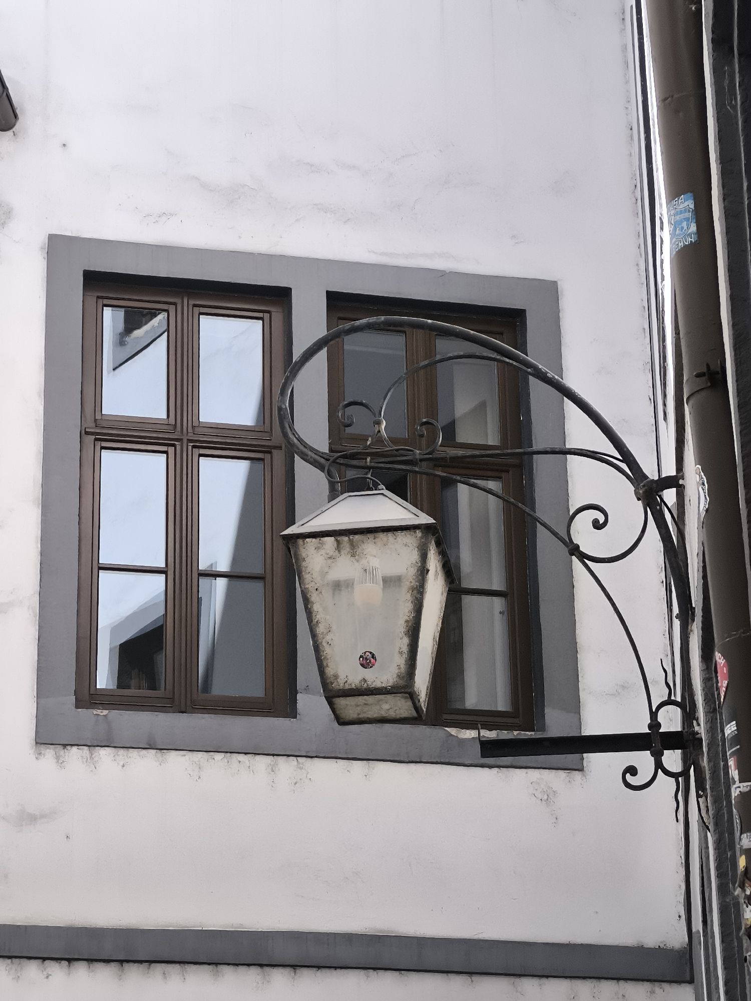 A close-up, low-angle, medium shot shows an outdoor lantern attached to a white, textured building facade in Bratislava, Slovakia. The lantern is a white, weathered metal structure with a peaked roof and glass panels, illuminated by a visible bulb. It is suspended from an ornate, black metal bracket with curlicue designs. To the left of the lantern is a rectangular window with a dark brown frame, divided into four panes by dark brown mullions. The window reflects the sky and surrounding buildings. To the right of the lantern and window, a portion of a downspout is visible, with a blue and white sticker partially attached. The building's facade is primarily white with some gray trim around the window and along horizontal bands. The scene appears to be on a street or alleyway.