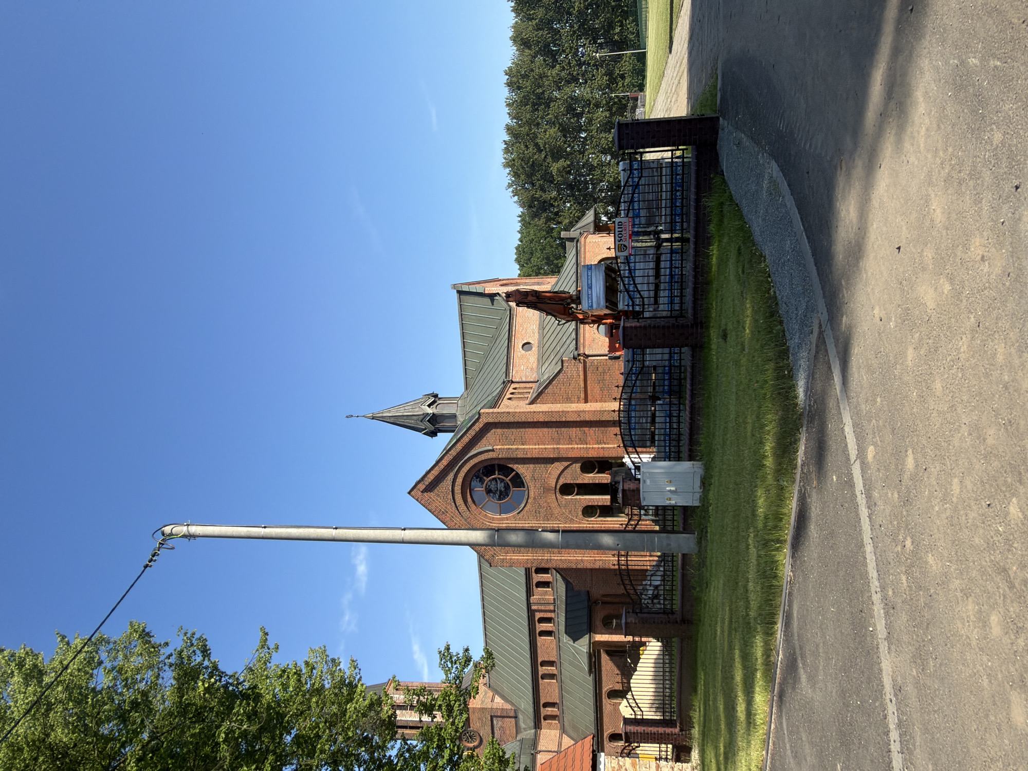 A brick church with a green roof and a steeple stands under a clear blue sky. A large tree with green leaves is on the left side of the image. A concrete utility pole with wires is in the foreground. In front of the church, there is a black metal fence, a patch of grass, and some construction materials, including an excavator and stacks of stone blocks. A sign on the stone blocks reads "SOLID SECURITY". The church is located in Stanowice, Poland.