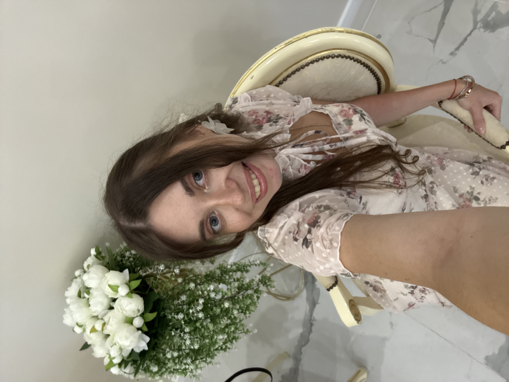 A young woman with long, dark brown hair and blue eyes is seated in a cream-colored chair with gold trim. She is wearing a light pink dress with a floral pattern and white polka dots, and she has white floral earrings. A large arrangement of white peonies and green foliage is visible behind and to her left. The floor is white marble. The woman is holding a phone in her right hand, as if taking a selfie, and is smiling. The background is a plain, light-colored wall.