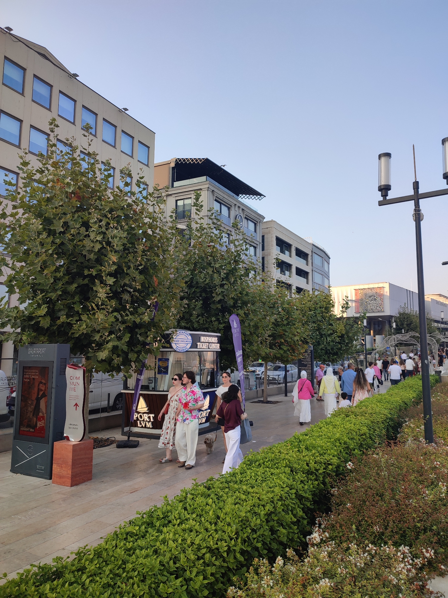 The image depicts a street scene in the city of Beyoğlu, Turkey. A group of people are walking along a paved walkway lined with lush green hedges. Several trees with large, leafy canopies are situated along the walkway. Modern buildings with large windows are visible in the background, suggesting a commercial or residential area. There is a ticket center labeled "BOSPHORUS TICKET CENTER" and a digital information stand. The sky is clear and blue. People are dressed in casual summer clothing, with some individuals wearing headscarves. The overall atmosphere appears to be a pleasant outdoor gathering or stroll.
