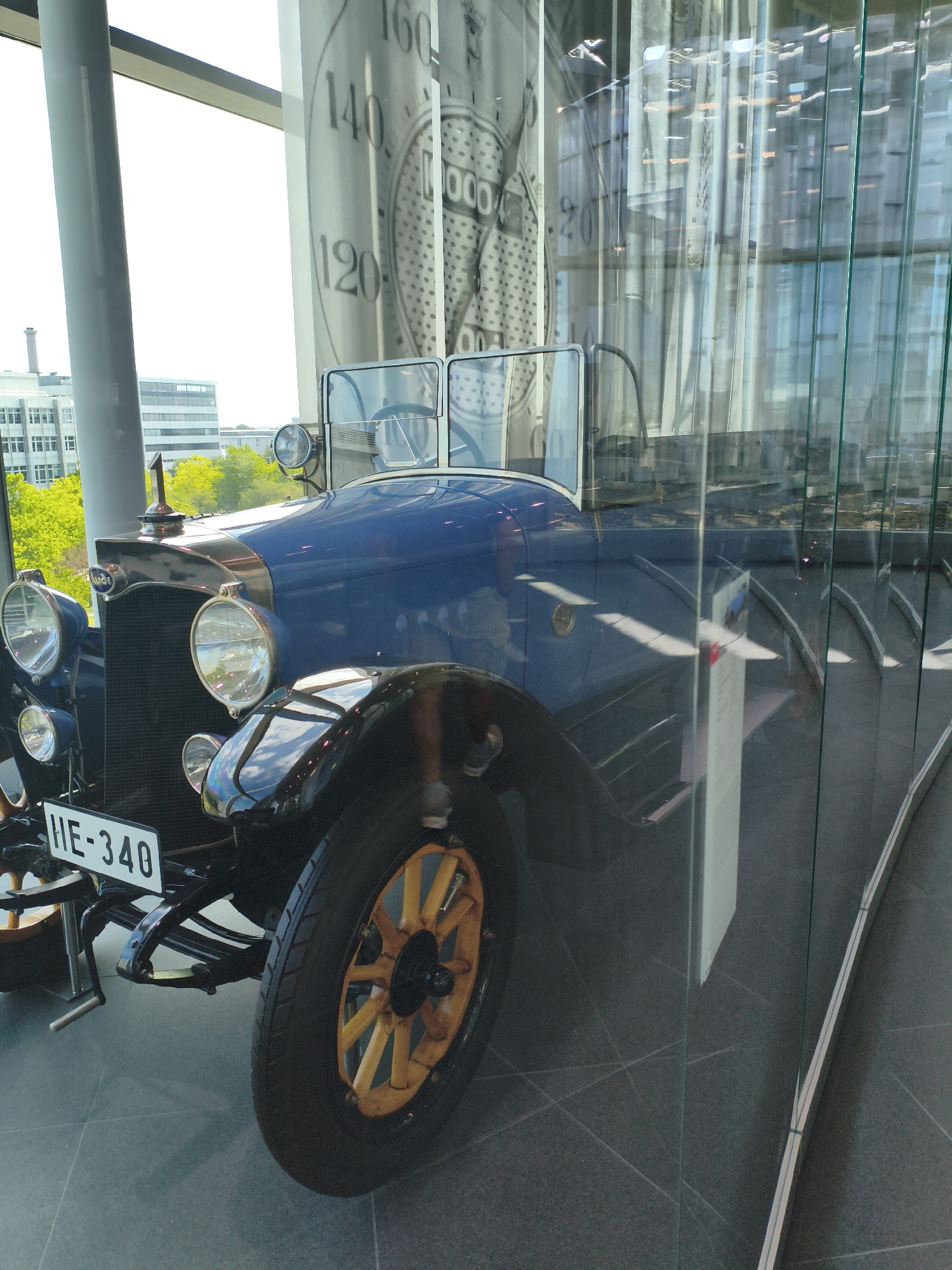 A vintage blue automobile is displayed behind glass, presumably in a museum, with its front quarter visible. The car has a dark grille, two large circular headlights, and a license plate reading "HE-340". The wheels have wooden spokes. In the background, through the glass and a large window, a modern city skyline with buildings and trees is visible. The floor is a dark, tiled surface. A large, meter-like graphic is visible on a fabric backdrop behind the car, featuring numbers and what appear to be speed readings. This scene is located at the Audi Museum Mobile in Ingolstadt, Germany.