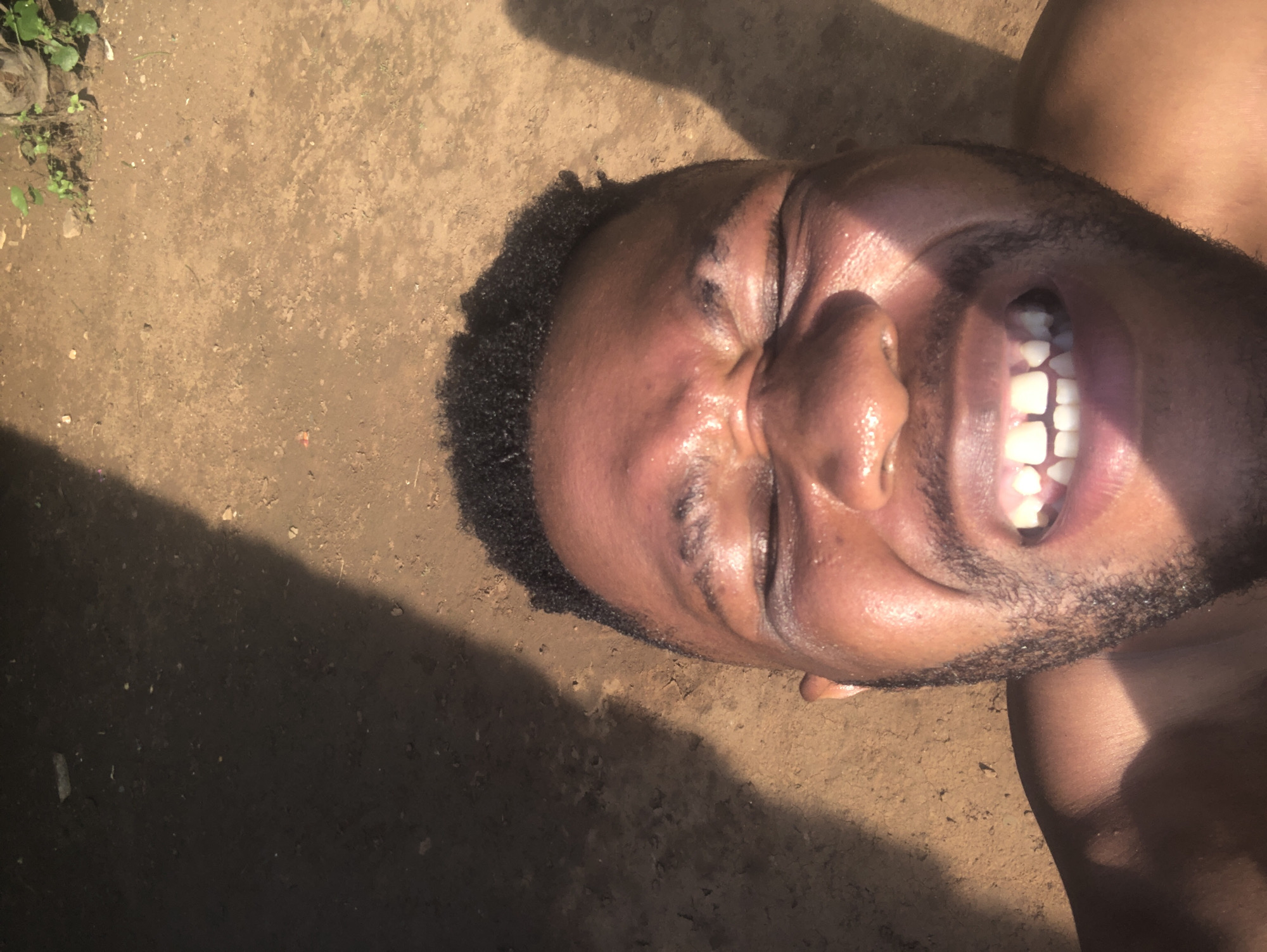 The image is a close-up, high-angle shot of a man's face and upper torso, rotated approximately 90 degrees clockwise, placing his head on the left and body extending to the right. The man has dark skin, short, curly black hair, and a neatly trimmed beard. He is smiling or laughing broadly, with his eyes squeezed shut and teeth prominently displayed. His skin appears shiny, possibly from sweat or oil, and is brightly illuminated by direct sunlight, creating strong highlights and deep shadows across his features, particularly on his forehead and nose.
The background consists of light brown, dry earth or dirt, suggesting an outdoor setting. Small green plants or weeds are visible in the top left corner of the frame. A large, dark shadow cuts diagonally across the bottom left portion of the image, indicating intense light from an unseen source, likely the sun. The strong sunlight and shadows suggest it is daytime, possibly midday or early afternoon. The man appears joyful or amused, perhaps reacting to the bright sun. There is no visible text in the image.