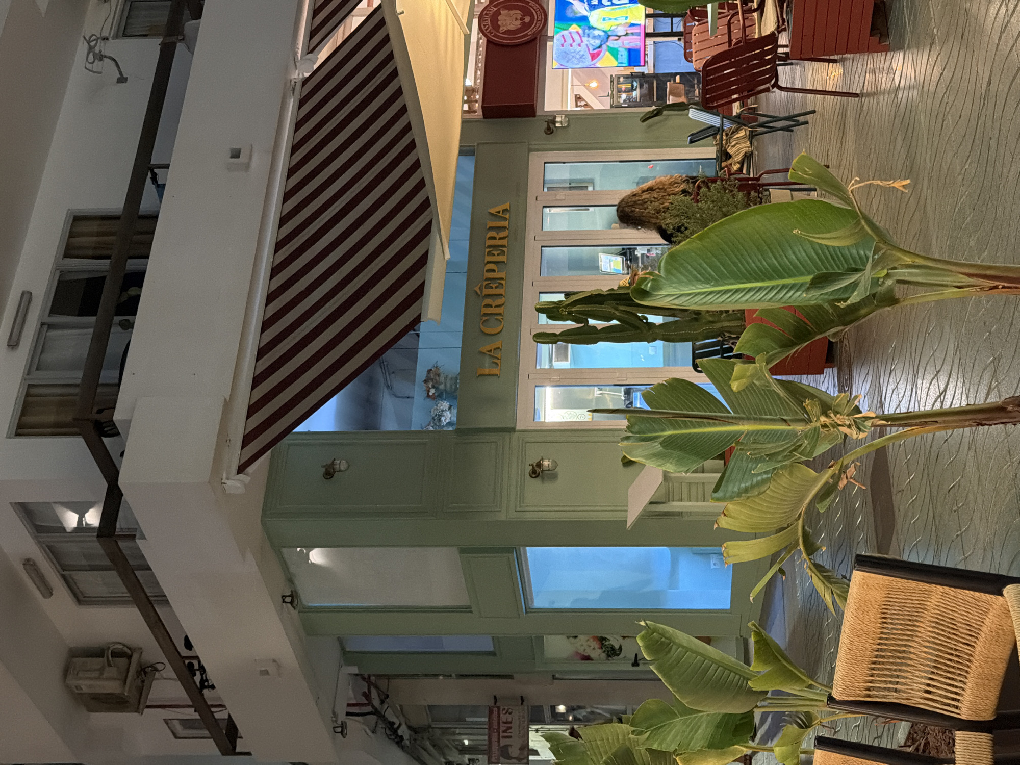 This image shows the exterior of "La Creperia," a crepe shop in Sant Antoni De Portmany, Spain. The shop has large glass doors and windows, with a red and white striped awning overhead. In front of the shop are several large potted plants, including a cactus. A person with curly blonde hair is sitting at a table inside the shop. The floor is paved with decorative tiles. There is a second story above the shop with a balcony and an air conditioning unit. To the left of the crepe shop, another business with a sign for "INES" is visible.