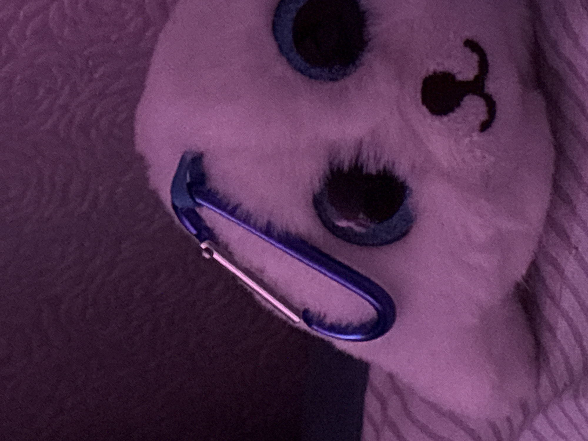 The image shows a white plush toy with blue eyes and a black stitched nose, resembling a bear. A blue carabiner clip is attached to the toy's fur. The background consists of a purple surface with a striped pattern. The image appears to be taken indoors under dim lighting conditions. The location in the city of Прикамский, Russia is not discernible from the image.