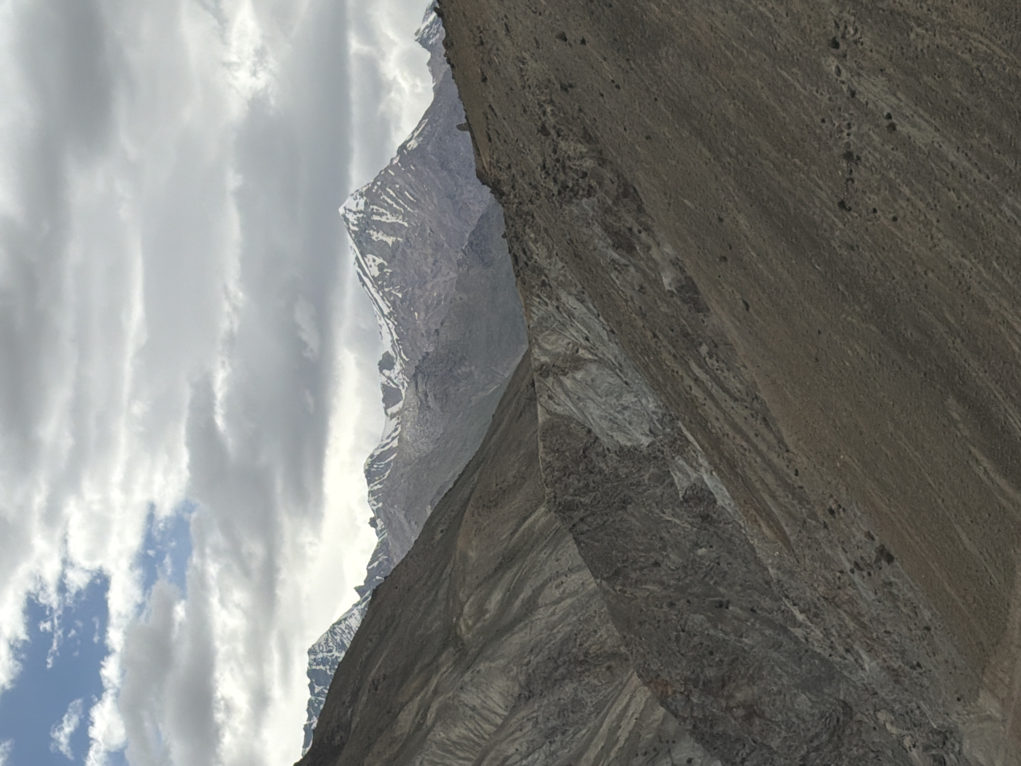 The image depicts a vast, mountainous landscape under a cloudy sky. The foreground is dominated by the rugged, barren slopes of mountains, characterized by dry earth, rocks, and visible erosion patterns. These slopes are devoid of vegetation. In the mid-ground, more mountainous terrain is visible, with a mix of rocky and scree-covered surfaces. The background features towering, snow-capped mountains, their peaks sharp and distinct against the overcast sky. The clouds are a mix of white and grey, with patches of blue sky peeking through in the upper left corner. The overall impression is one of a remote, high-altitude environment, characteristic of regions like the Himalayas in India, potentially near areas like Testa, known for its dramatic mountainous geography. There are no people or man-made structures clearly visible in the image.
