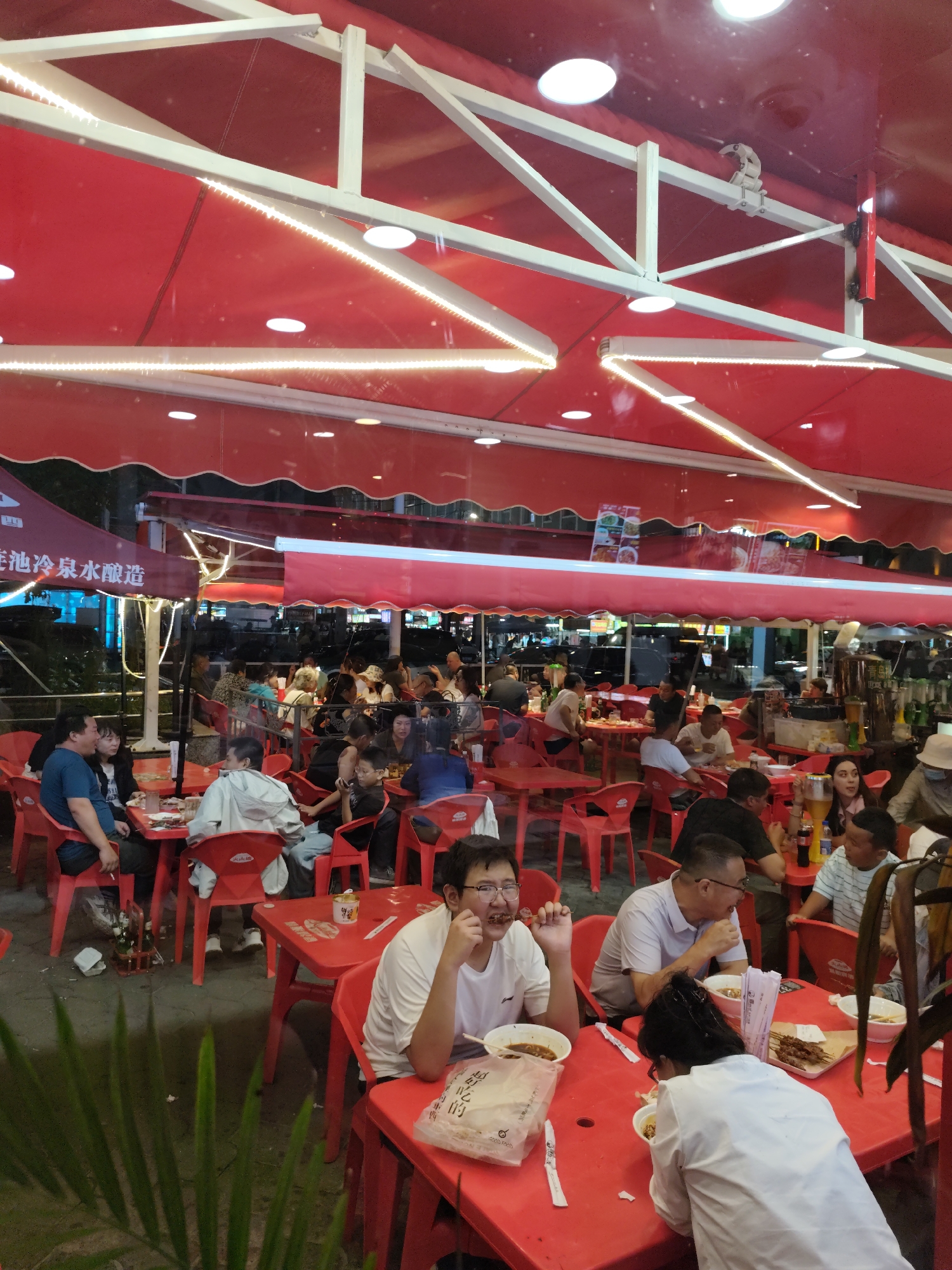 This image depicts an outdoor dining scene at a restaurant in Aihui, China. Numerous patrons are seated at red plastic tables and chairs under a large red awning supported by white metal structures. The awning is illuminated by string lights and circular light fixtures. The ground appears to be paved, and a potted plant with large green leaves is visible in the foreground.
Several individuals are engaged in eating and conversation. In the foreground, a young man wearing a white shirt with glasses is actively eating from a bowl of food with chopsticks. To his right, another person in a white shirt is looking down at their food. In the middle ground, a group of people are seated around a table. A man in a blue shirt is seen on the left side of the image, conversing with a woman. Further back, more tables are occupied by diners, some of whom are holding drinks and food.
The overall atmosphere suggests a lively and casual dining environment. The presence of food, drinks, and social interaction among the patrons indicates a typical restaurant setting. The red color scheme of the awning and furniture creates a vibrant and cohesive visual theme. The background shows the urban environment with blurred lights and structures, suggesting the restaurant is situated in a populated area of Aihui.