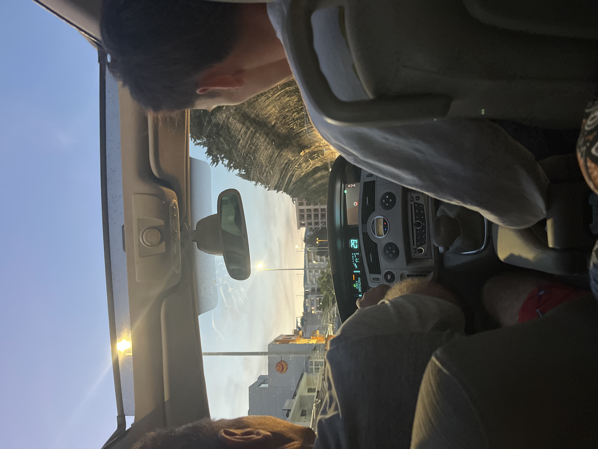 The image is a point-of-view shot from inside a car, looking forward. Two individuals are visible. In the foreground, a person is seated in the driver's seat, their back to the camera, wearing a grey shirt and red shorts. To their right, another person is in the passenger seat, also facing forward with their back to the camera, wearing a light-colored long-sleeved shirt. The car has a sunroof through which the sky is visible. The dashboard of the car is in the center of the frame, displaying various controls and a digital readout. Outside the car, a cityscape with buildings and streetlights can be seen, bathed in the warm light of either sunrise or sunset. The overall impression is of a drive through a town during twilight. The location is identified as Pistula, Montenegro.
