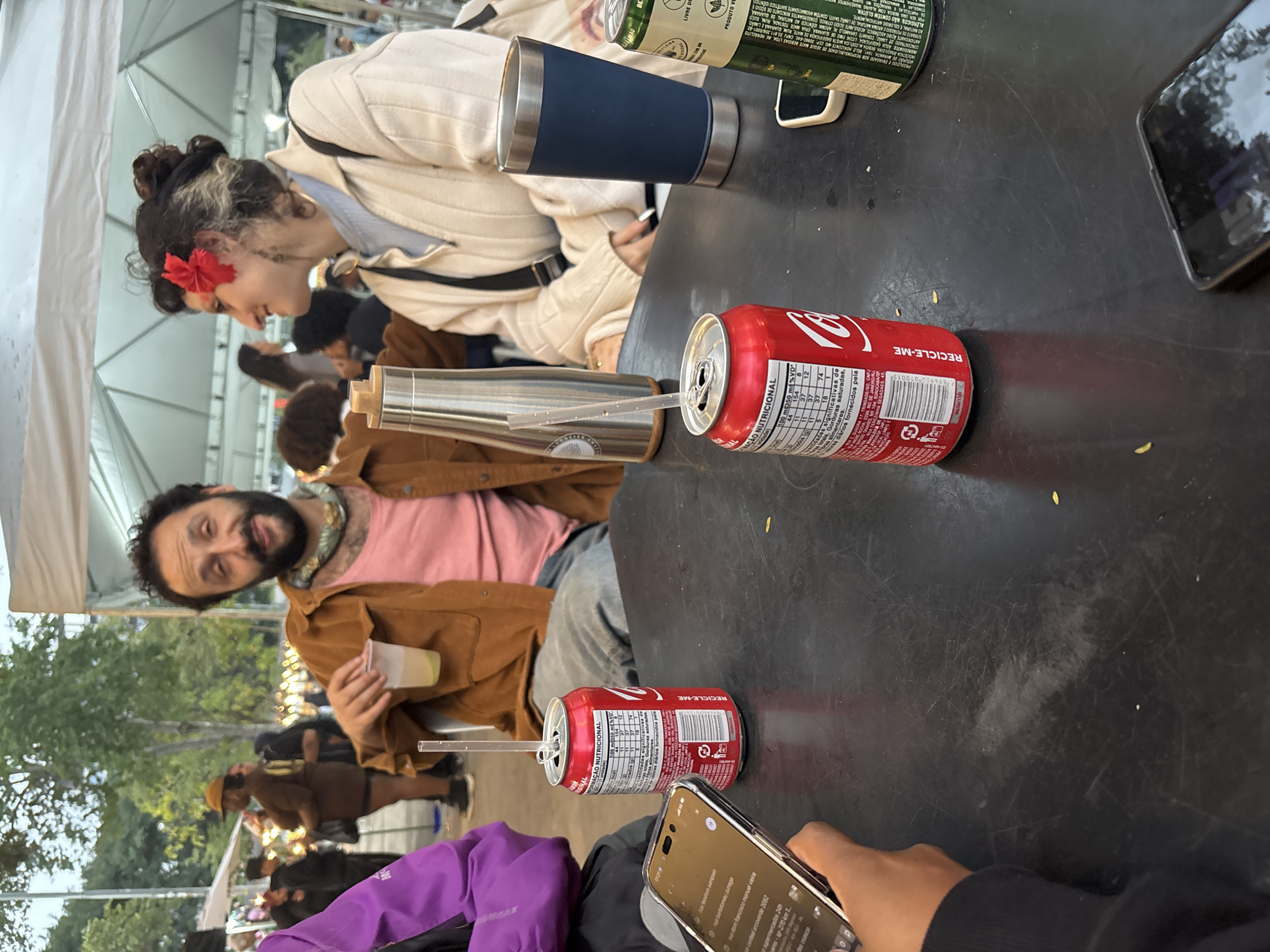 The image shows a group of people sitting around a black table. On the table are two open cans of Coca-Cola with straws, a stainless steel thermos, a blue tumbler, a green and white beverage container, and two smartphones. One person is holding a clear plastic cup filled with a light-colored beverage. Another person is wearing a red flower in their hair. The location appears to be outdoors at an event, with trees and other people visible in the background. Based on the OCR text found on the Coca-Cola cans, the location is São Paulo, Brazil.
