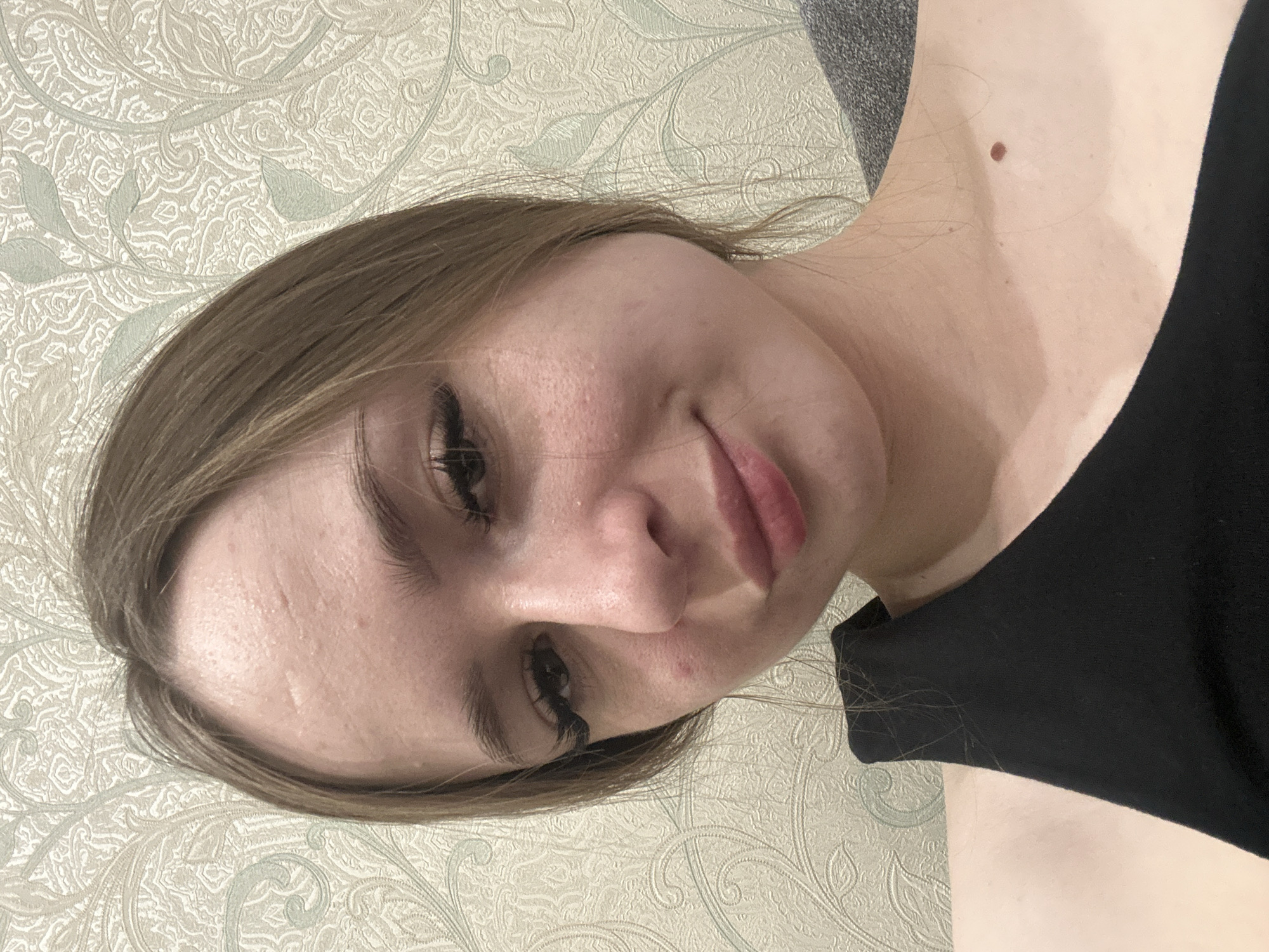 A close-up portrait depicts a light-skinned woman with brown hair, possibly indoors. She is wearing a black top, and a mole is visible on her neck. The background features a patterned wall. The location and city are indeterminate.