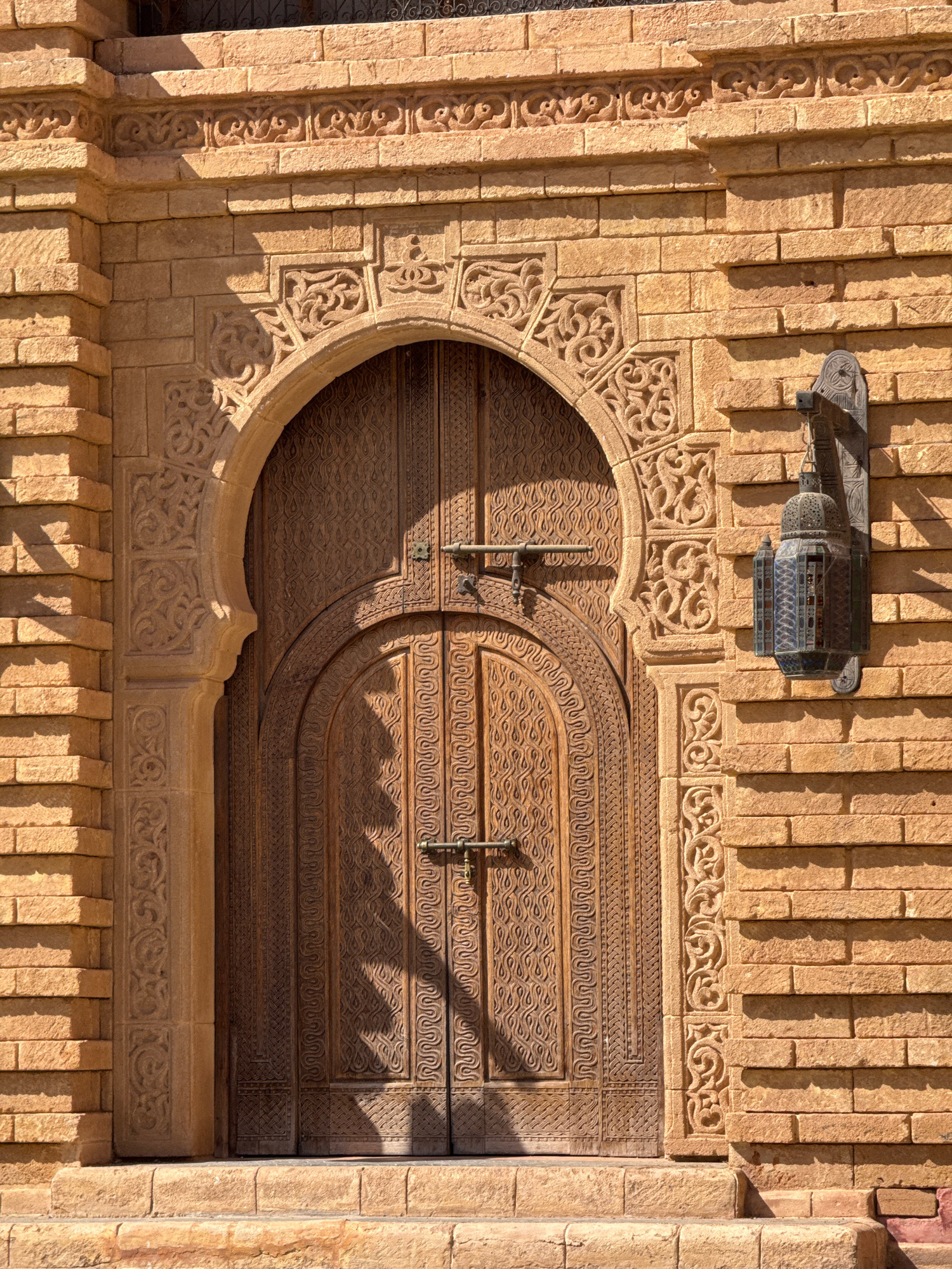 A traditional wooden door with intricate carvings is set into a sandstone brick wall. The door is arched at the top, with detailed carvings adorning the surrounding stone archway and pilasters. A wrought-iron lantern hangs on the right side of the wall. The scene appears to be an exterior architectural detail, likely from a historical building in Agadir, Morocco, given the style of the architecture and ornamentation. The lighting suggests it is daytime, with sunlight casting shadows on the door and wall. There are no people or animals visible in the image.