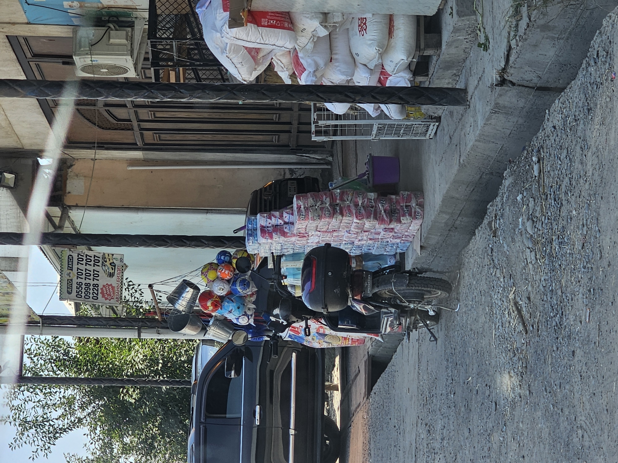 This is a street-level, medium shot photograph taken in the city of Özgön, Kyrgyzstan. The image captures a street scene with various commercial elements. In the foreground, a dark-colored electric scooter is parked, facing away from the viewer. It has a black storage box on the back and a license plate that is partially visible. To the left of the scooter, a black SUV is parked.
Adjacent to the scooter, a collection of colorful balls, possibly for sale, are hanging from a rack, along with several metal buckets. A sign, partially obscured by the scooter and balls, hangs on a pole, displaying phone numbers and text in Cyrillic and Latin scripts, indicating a taxi service. In the background, stacks of packaged goods, likely hygiene products or food items, are arranged on the pavement. Further back, a building with wooden doors and an air conditioning unit mounted on the exterior wall is visible. The ground is covered with gravel and dirt, and a concrete drainage ditch runs along the edge of the sidewalk. The scene is illuminated by natural daylight, casting shadows and highlighting the textures of the objects and ground.