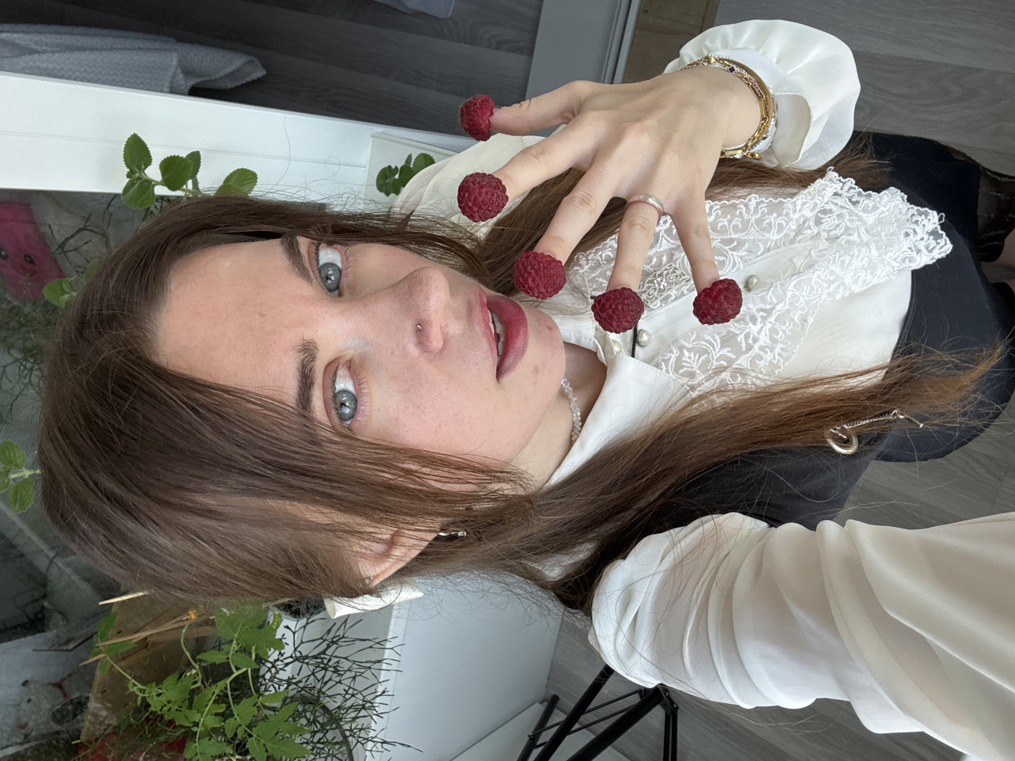 An adult woman with medium-length brown hair and blue eyes is shown from the chest up, looking into a camera at a slightly upward angle. She is wearing a white collared blouse with lace detailing and pearl buttons, a black vest or dress over it, and a delicate necklace. Her left hand is raised, with raspberries placed on the tips of her index, middle, ring, and pinky fingers. She is also wearing a silver ring on her left ring finger and a gold bracelet on her left wrist. The background appears to be an indoor setting, possibly a room with plants visible.