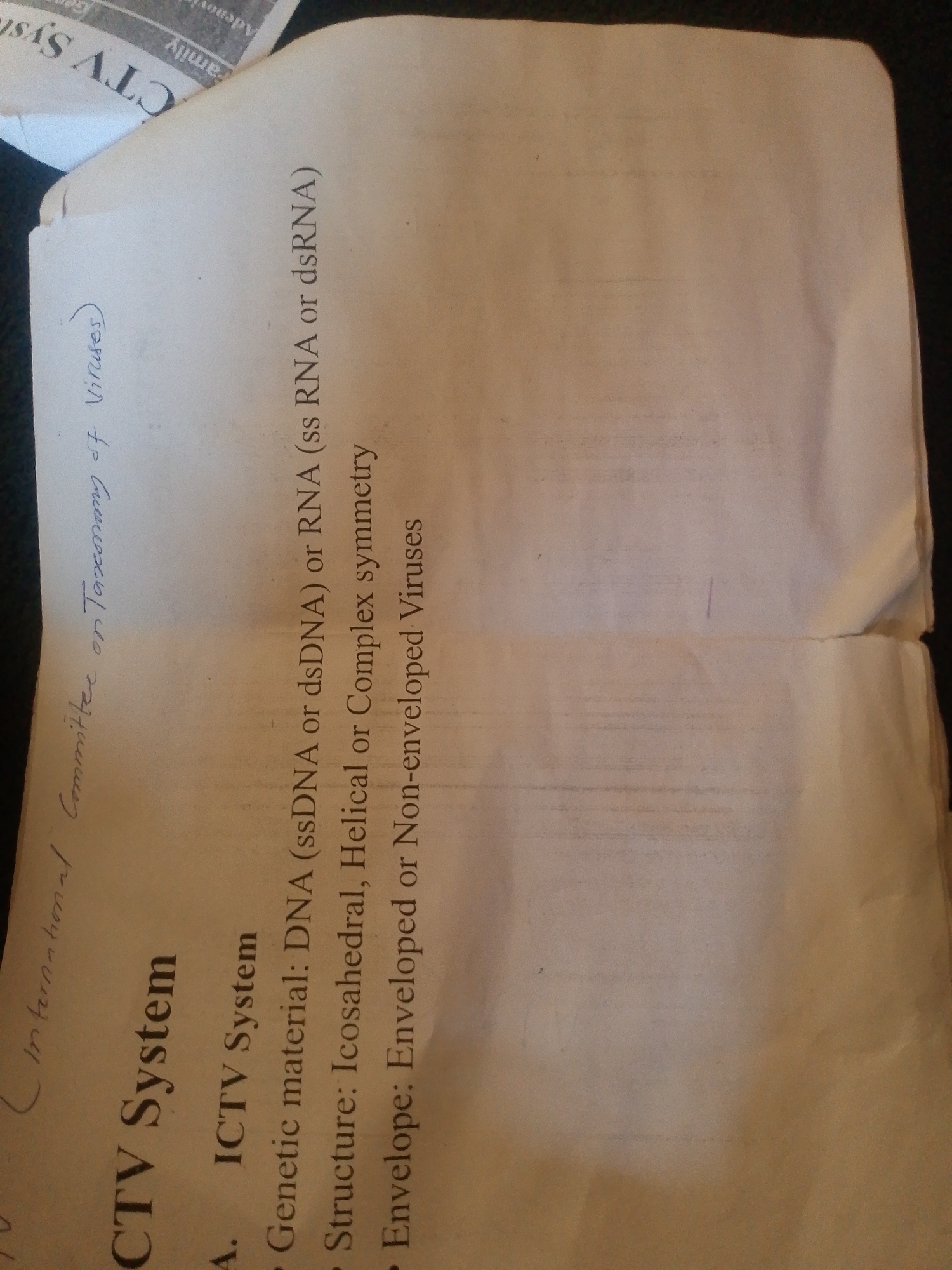 The image shows a close-up view of a document with text printed and handwritten on it. The document is lightly creased and appears to be part of a larger stack of papers. The text is oriented vertically in the image, suggesting the original document was held or placed in a portrait orientation.
Printed text includes headings like "ICTV System" and bullet points detailing characteristics such as "Genetic material: DNA (ssDNA or dsDNA) or RNA (ss RNA or dsRNA)", "Structure: Icosahedral, Helical or Complex symmetry", and "Envelope: Enveloped or Non-enveloped Viruses".
Handwritten text, in a different style and color, is visible in the top left and along the left edge, reading "International Committee on Taxonomy of Viruses". There is also some faint, possibly printed, text at the very top that is mostly cut off and illegible, but appears to be part of a newspaper clipping or a different document layered beneath.
There are no people, animals, or any other elements indicative of a specific location like Jalingo, Nigeria. The scene is entirely focused on the surface of the paper and its textual content. The lighting suggests an indoor environment, likely with overhead or ambient light. The background is dark, indicating it is likely a surface the document is resting on.