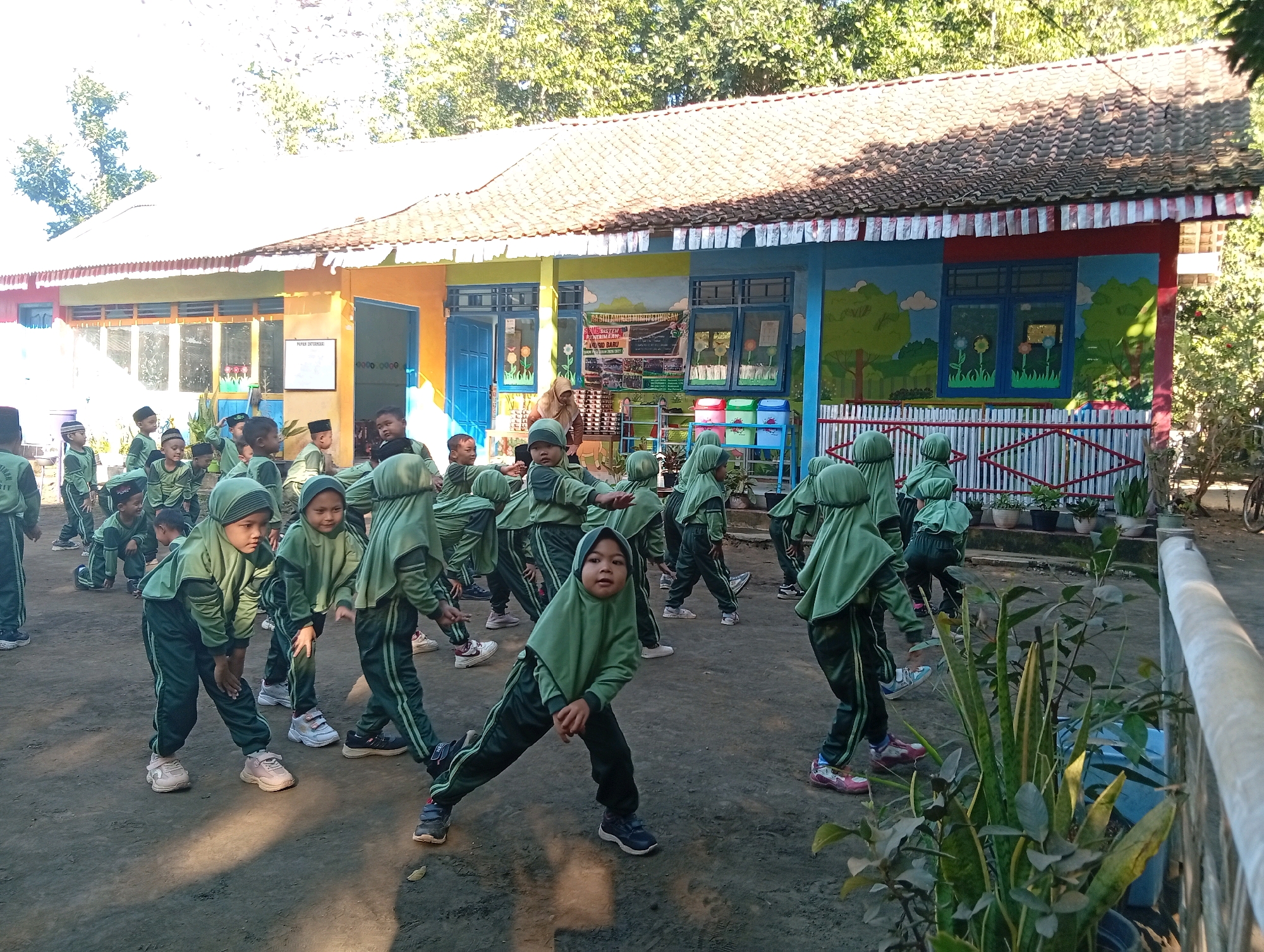 A group of young children, likely students, are engaged in an outdoor physical activity in front of a building with a red-tiled roof. The children are all wearing matching green uniforms with headscarves and dark pants. They appear to be performing some kind of stretching or exercise, with several children in lunging or bent-over poses. The building behind them is brightly colored with murals depicting trees and clouds. There is a fence in front of the building, and potted plants are visible. The ground is dirt and gravel, suggesting a schoolyard or similar outdoor area. The location is identified as Miritpetikusan, Indonesia.
