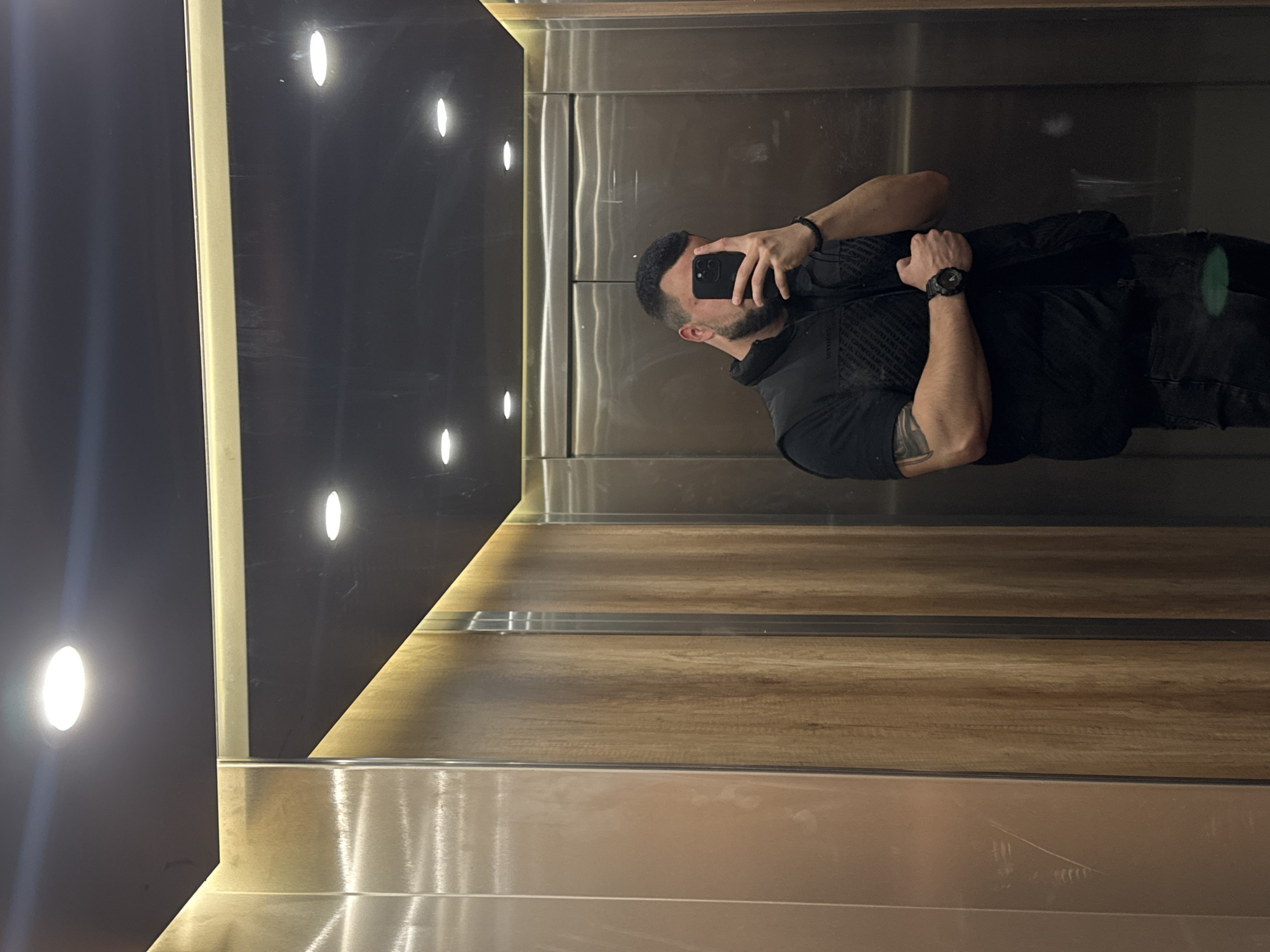 A man is taking a selfie in an elevator with interior walls of dark, reflective material and a light wood paneling section, situated in Sofia, Bulgaria. The elevator has multiple bright ceiling lights. The man is wearing black clothing, including a watch, and has a beard and visible tattoos on his arm. He is holding a smartphone to take the photo. The elevator interior appears modern and well-maintained.