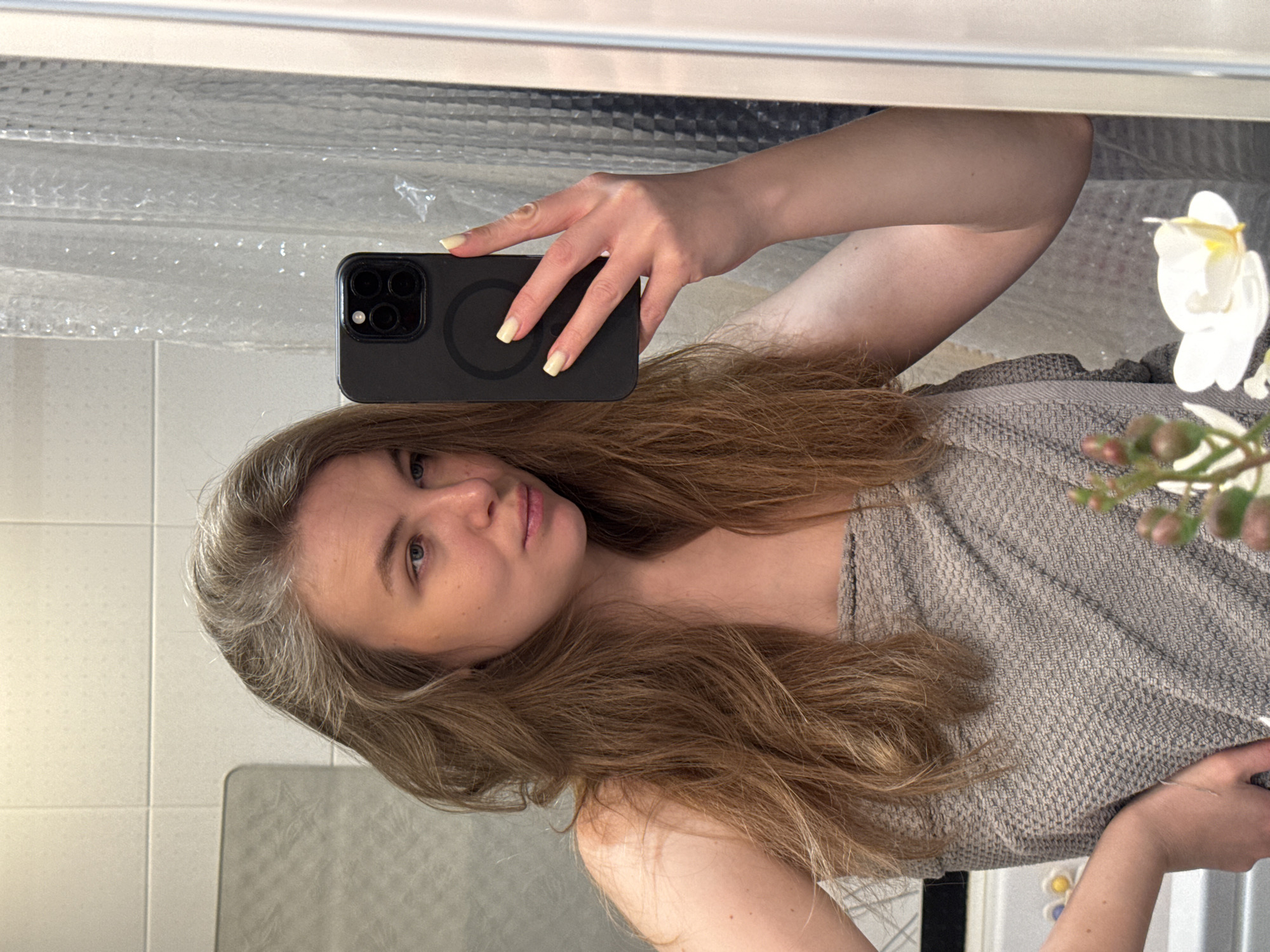 A person is taking a selfie in a bathroom with white tiled walls and a mirror. The person is holding a black smartphone in their left hand, partially obscuring their face. They have light brown hair with some grey roots visible. They are wearing a grey top. A white artificial flower is visible in the background to the right. The image is taken indoors. The location is likely a private residence. There is no evidence to indicate the location is Vladimir, Russia.