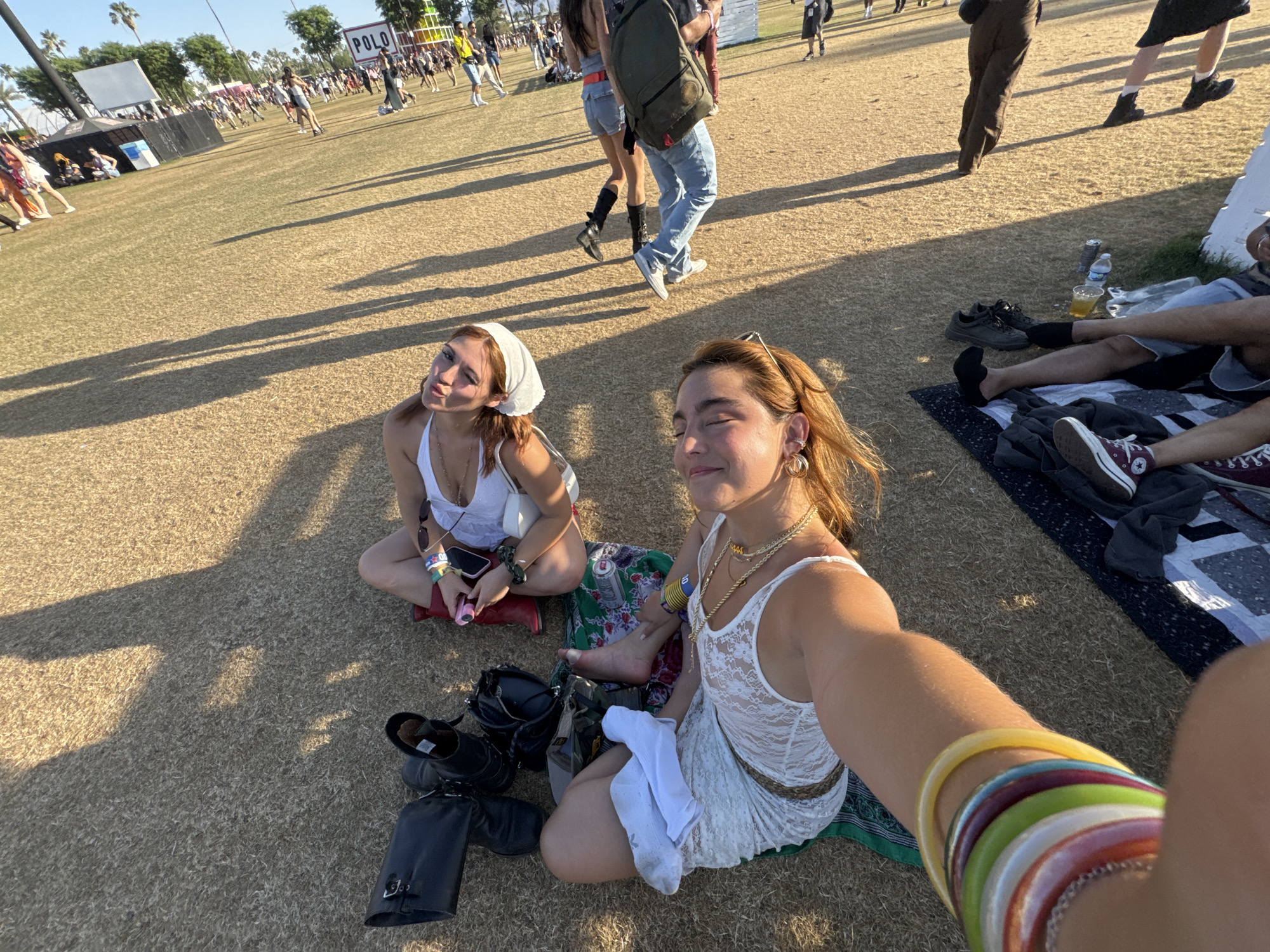 The image captures a sunny day at a grassy outdoor venue, likely the Empire Polo Club in Indio, California, United States, given the 'POLO' sign in the background. Two young women are seated on a patterned blanket in the foreground. The woman on the left has auburn hair covered by a white headscarf and wears a white top. The woman on the right, who appears to be taking the selfie, has shoulder-length auburn hair, gold jewelry, and a white lace dress. A collection of colorful bracelets adorn her outstretched arm. Their belongings, including boots, a black bag, and a can of soda, are scattered around them. In the background, people are walking and socializing on the grass, suggesting a large gathering or festival. Further back is a stage or structure and rows of people.