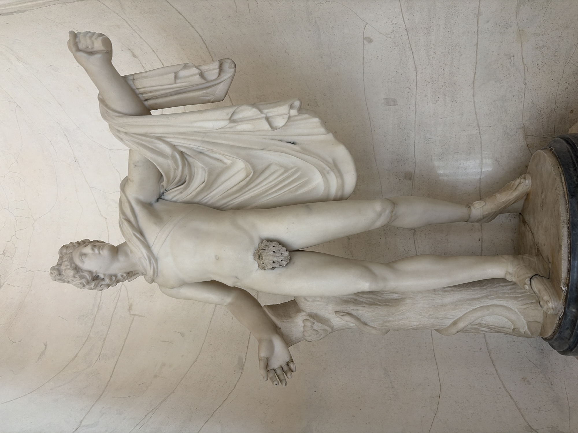 The image depicts a marble statue of a male figure. The statue is lying on its side. The figure is nude, partially draped with a cloth. His right arm is raised with a clenched fist. His hair is styled in tight curls. The statue rests on a cylindrical base. The background consists of a wall with noticeable cracks. The statue is located in the city of Unknown, Unknown.