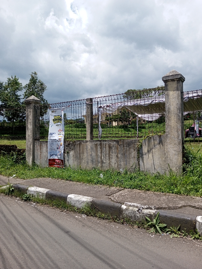 The image depicts an outdoor scene in Bagochen Boolingpang Ueling, Bhutan, under a heavily overcast sky. In the immediate foreground, a grey asphalt road is visible on the left, bordered by a black and white painted concrete curb. A strip of unkempt grass and weeds grows between the curb and a perimeter fence.
The midground is dominated by a fence composed of three visible rough concrete pillars with pyramidal caps, supporting a dark wire mesh. A lower concrete wall forms the base of this fence. A tall, vertical white banner with colorful graphics and text, including "BAZAAR RAMADHAN" and "VOTE?", is affixed to the wire mesh between the two leftmost concrete pillars. Overgrown green vegetation, consisting of grass and weeds, is present around the fence line on both sides of the concrete wall.
Behind the fence, in the background, a light-colored, multi-story building with a brown roof is visible, partially obscured by trees and several white, tarp-like temporary structures or shelters. Additional trees are visible further to the left, and the ground in the background is green and unmanicured. No individuals or active movements are present in the scene.