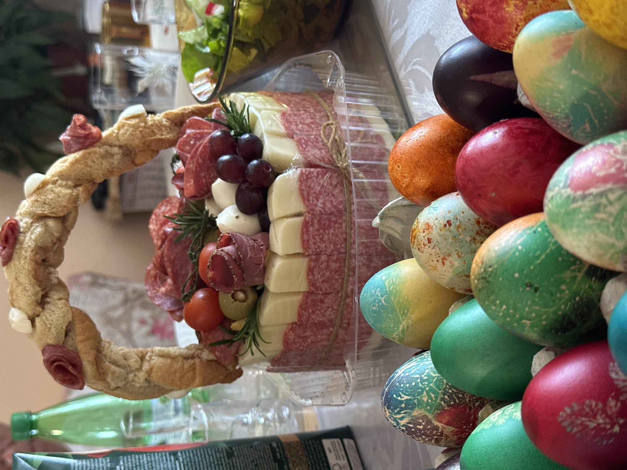 On a table in Rakovski, Bulgaria, sits a charcuterie arrangement in a clear plastic container, featuring stacks of sliced cheese and salami tied with twine. Grapes, mozzarella balls, tomatoes, olives, and rosemary garnish the arrangement. A bread wreath with salami rosettes encircles the charcuterie. A pile of colored Easter eggs sits to the right of the charcuterie. A salad in a clear bowl is visible in the background.