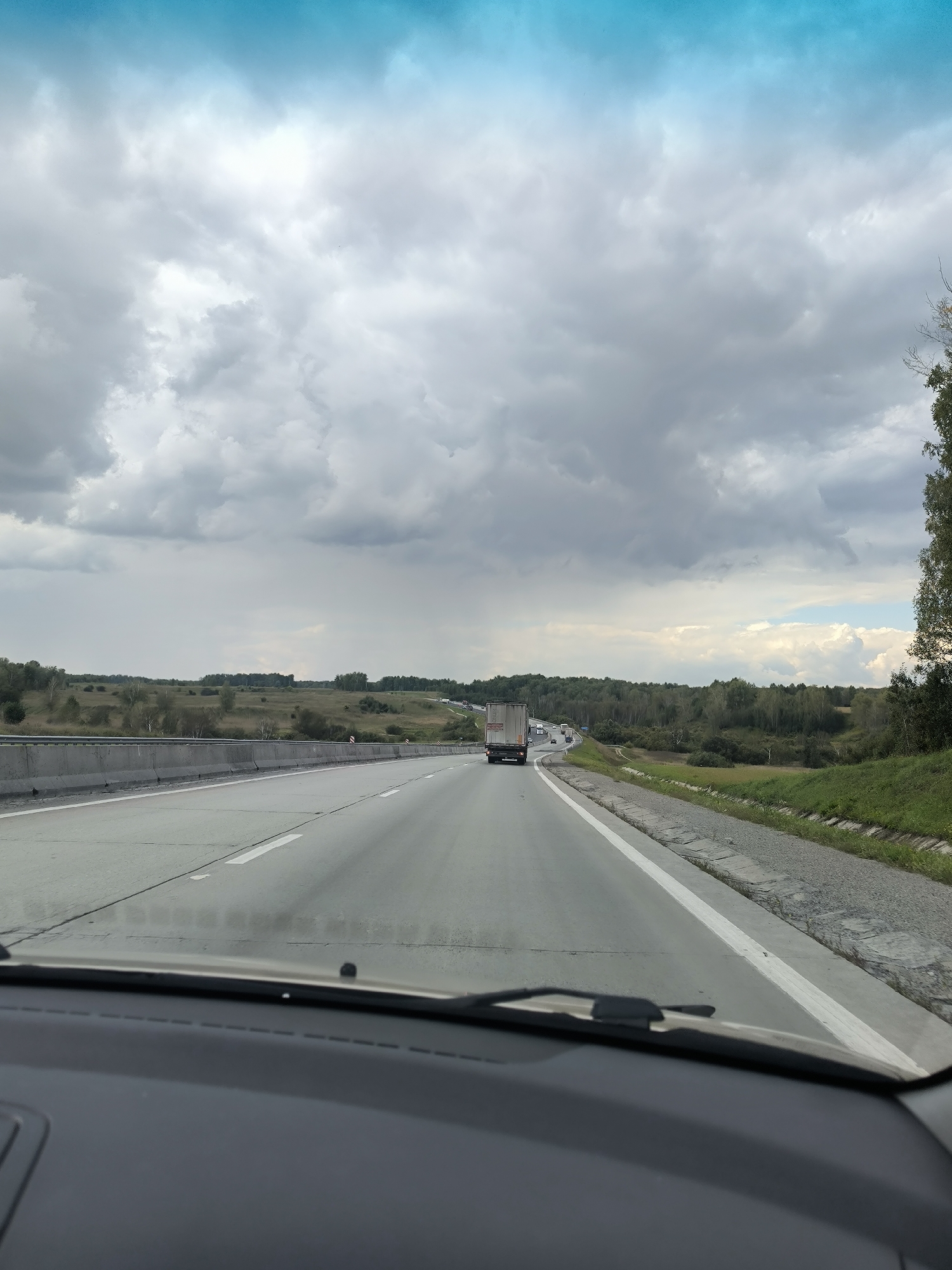 The image is a first-person view from a vehicle traveling on a highway. The sky is overcast with dramatic, gray clouds, but a bright blue patch is visible near the top. The highway stretches ahead, with multiple lanes and a concrete barrier on the left. A white truck is visible in the distance on the right side of the road, and other vehicles are scattered further down the highway. The landscape on either side of the road consists of rolling hills covered in trees and fields, suggesting a rural or semi-rural setting. The foreground shows the dashboard and windshield wipers of the vehicle, indicating the perspective of the photographer. The overall impression is one of a journey on a highway under a moody sky, with the vastness of the landscape as a backdrop.