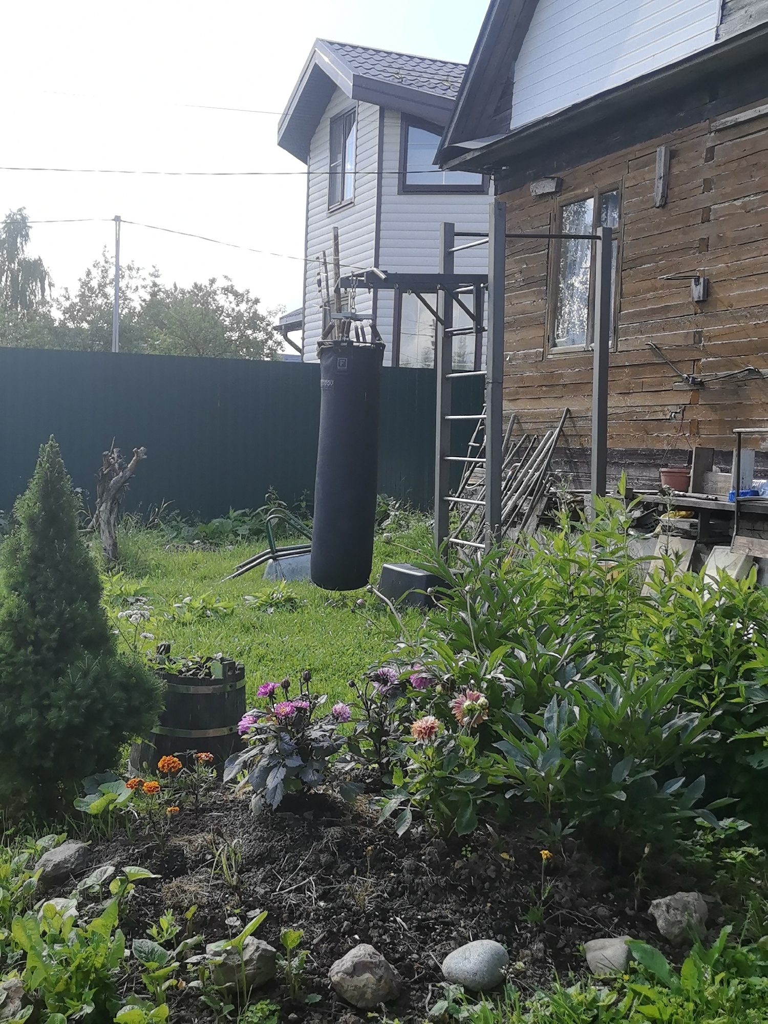 The image depicts an outdoor scene with a focus on a garden and a wooden house. In the foreground, there is a flowerbed with various plants and flowers, including orange marigolds and purple dahlias, alongside a small evergreen tree and a dark wooden barrel. The ground is covered with dark soil and some rocks. Behind the flowerbed, a punching bag is suspended from a metal frame attached to a rustic wooden building. This building appears weathered, with exposed wooden planks and a visible window. To the left of the wooden building is a newer, multi-story structure with white siding and a dark roof. The background consists of a dark green fence, a patch of grass, and trees. There are no people visible in the image. The scene is consistent with a residential yard, possibly in a rural or suburban setting. Given the information provided, the location is identified as Matchino, Russia.