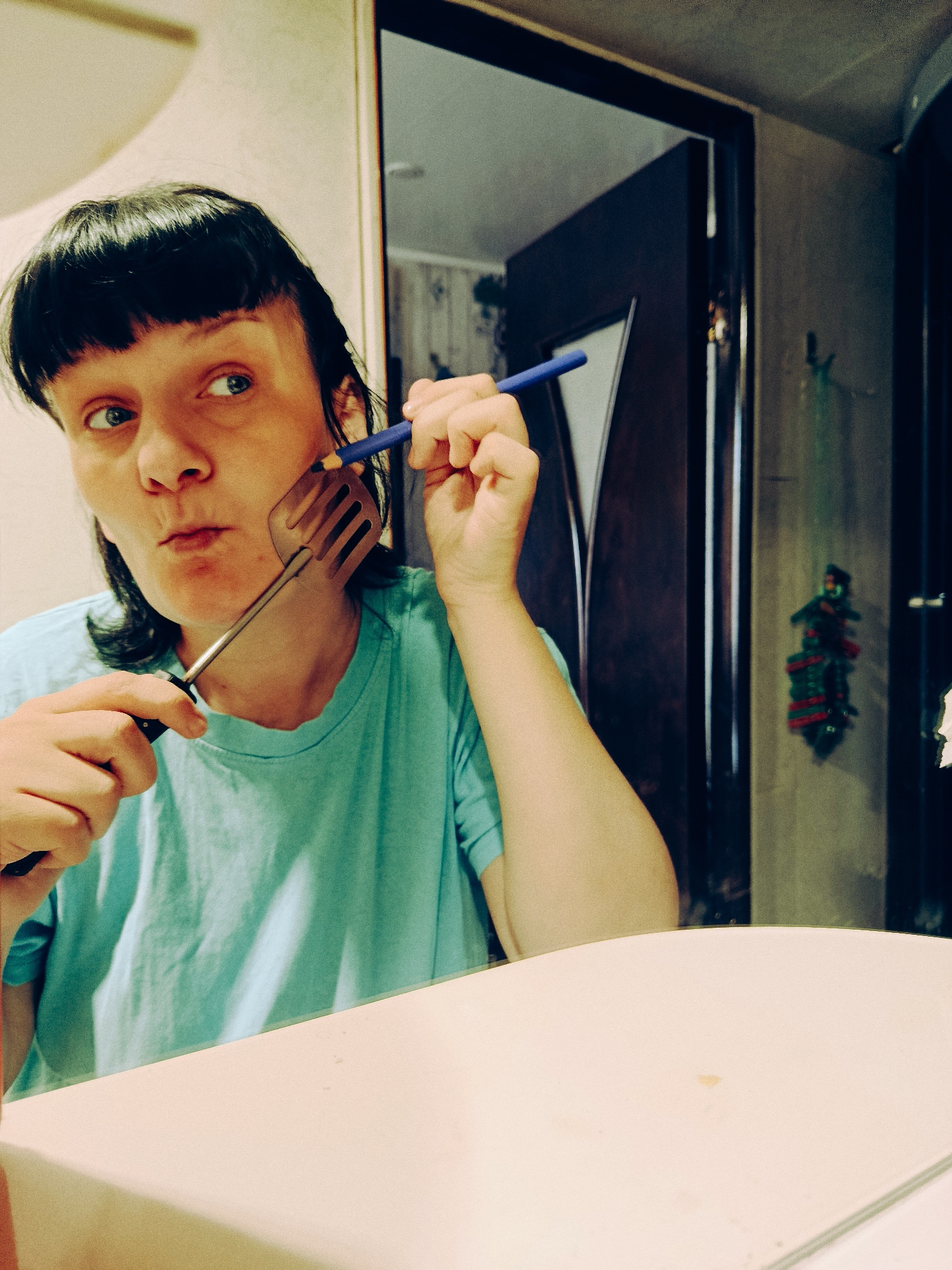 A person is holding a spatula to their face while holding a pencil in the other hand, as if shaving. The person has dark hair and is wearing a blue t-shirt. The scene appears to be a bathroom with a mirror and a door in the background. A decorative item hangs on the wall next to the door. The location is Фоки, Russia.