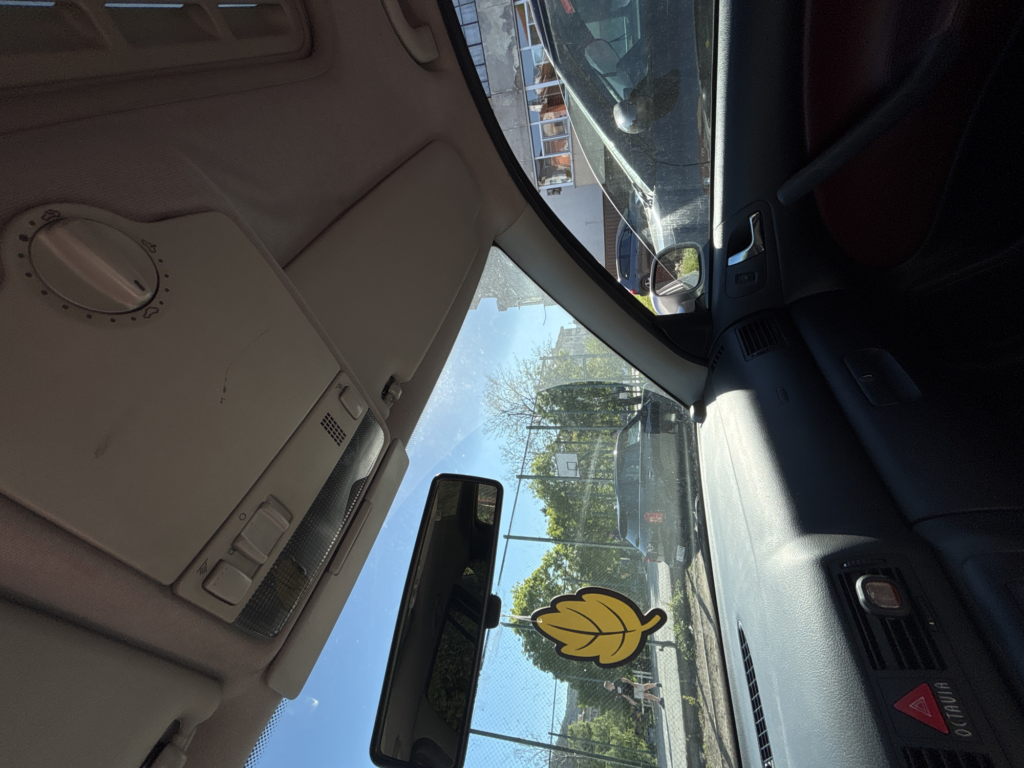 The image is taken from inside a car, looking out through the windshield. A yellow leaf-shaped air freshener is attached to the windshield. The car is parked on a street next to a sidewalk. On the sidewalk, two people are visible walking. In the background, there is a fenced area with trees and a basketball hoop. Buildings are visible beyond the trees. The make of the car is apparent on the dashboard as "OCTAVIA". The location is Unknown, Unknown.