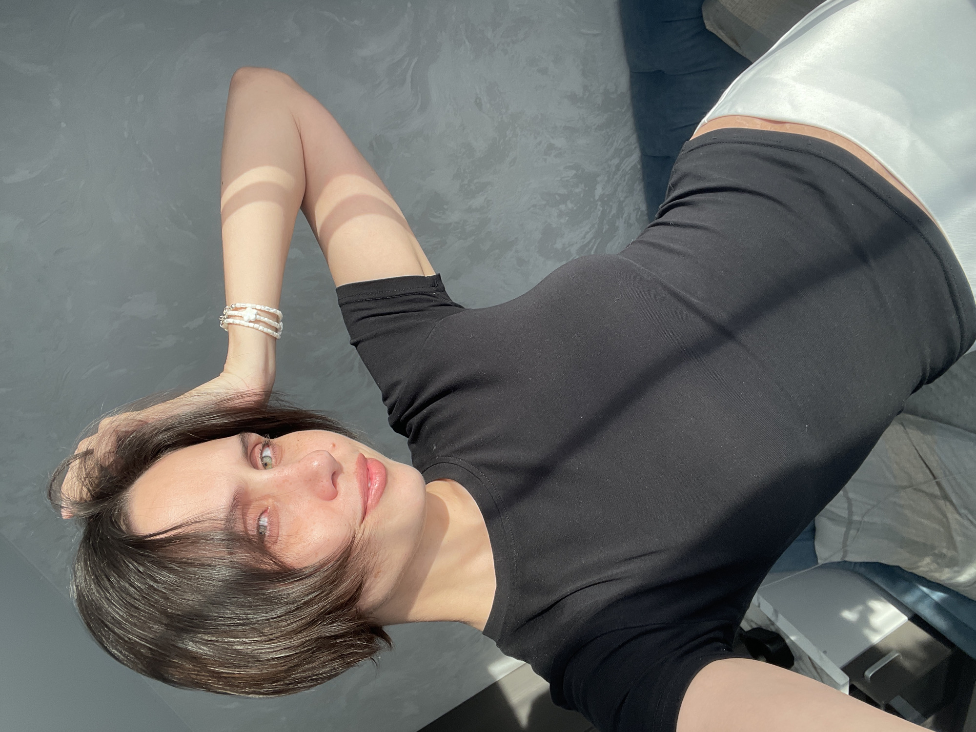A woman with short, dark hair and fair skin is lying down, facing the camera. She is wearing a black top and white pants. Her left arm is raised, and she is wearing a bracelet on her wrist. The background appears to be a textured gray wall, and part of a blue couch is visible on the right. While the image does not contain any information to definitively place the scene in Izhevsk, Russia, the photo was likely taken indoors.