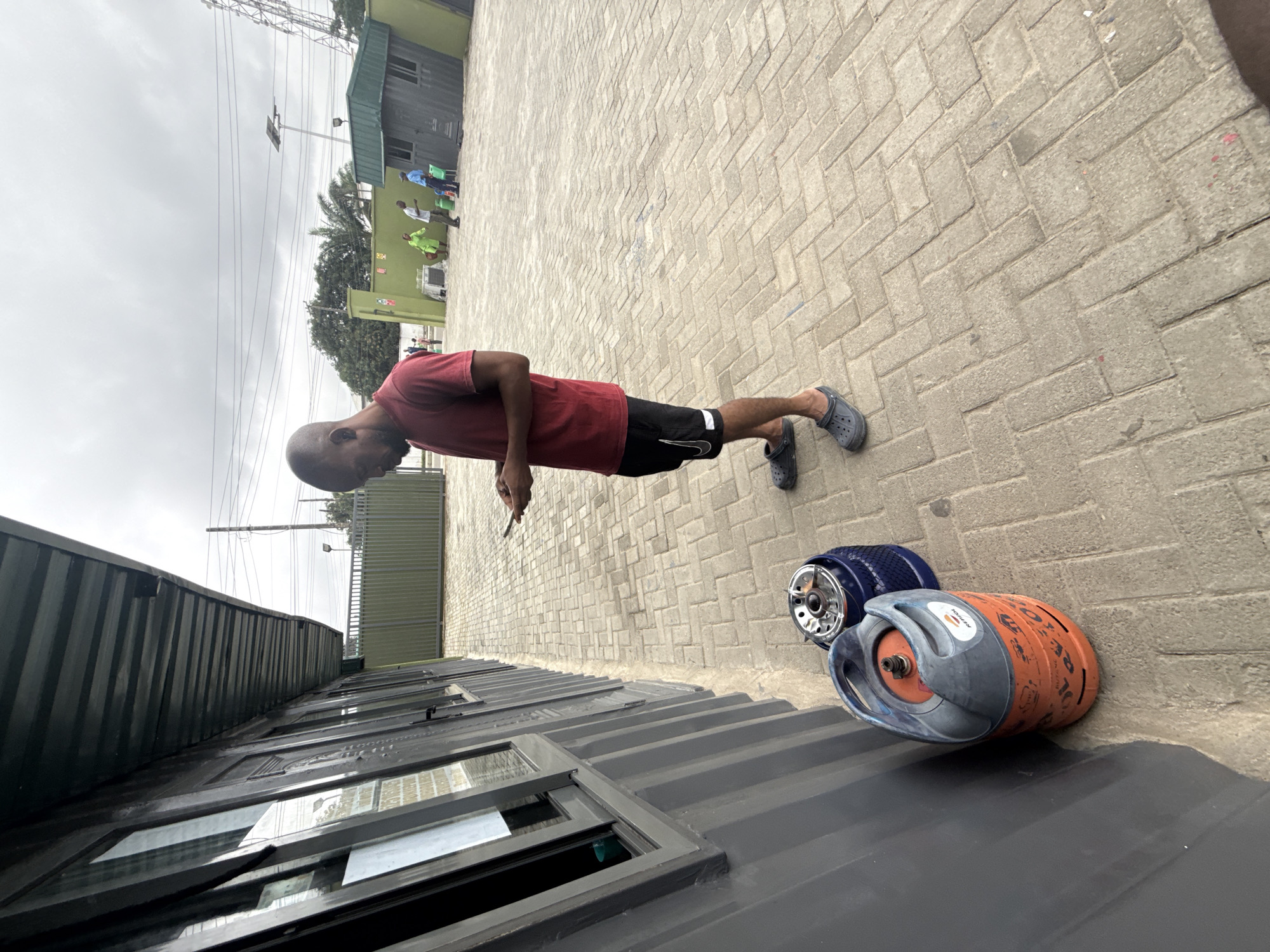 A man stands on a paved courtyard in front of a dark gray, corrugated building with multiple windows. He is wearing a red t-shirt and black shorts with white accents. He is looking down at his mobile phone, holding it with both hands. To his left, leaning against the building, are two gas cylinders: a larger orange one with "REPSOL" branding and a smaller blue one. The courtyard is paved with interlocking brick pavers. In the background, further down the courtyard, a green fence encloses an area with several people, a small green building, and what appears to be an air conditioning unit. The sky is overcast. This scene is likely located in Ado, Nigeria.