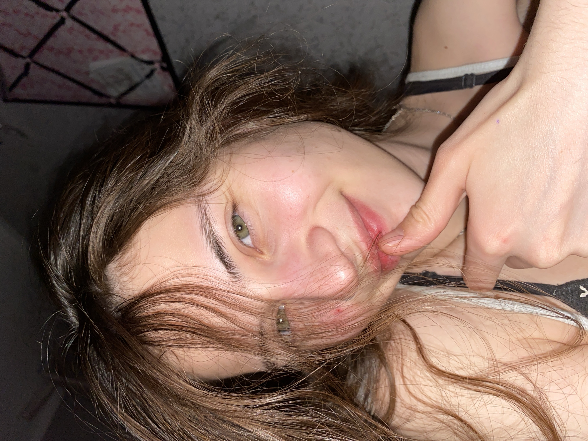 A young woman with light skin and brown hair is lying down, facing the camera. Her right hand is raised, with the index finger near her mouth. She has green eyes and is wearing a white and black bra or top. A black and white patterned object is visible in the upper left corner of the frame. A choker and necklace are also visible. The location is not discernible from the image. The location is not in Votkinsk, Russia.