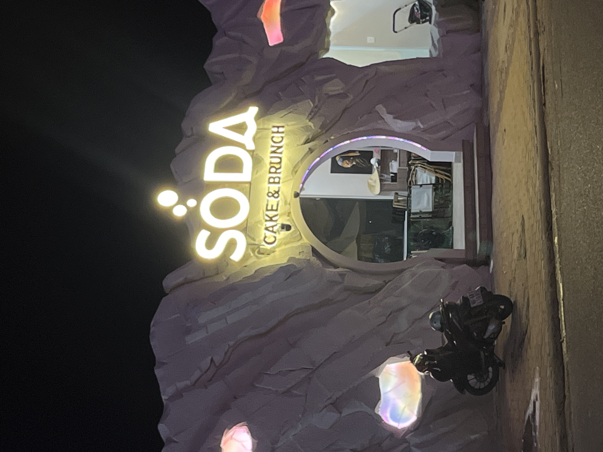 An exterior shot at night shows a building with a facade designed to resemble a rocky cave. The word "SODA" is illuminated in large, bright yellow letters above an arched entrance. Below "SODA," the text "CAKE & BRUNCH" is also visible, in smaller yellow letters. The entrance itself is an arched opening with purple LED lights outlining its edge.
Inside the entrance, a portion of the interior is visible, including a framed picture of "Girl with a Pearl Earring" by Johannes Vermeer hanging on the wall, and some furniture. A straw hat is placed on a table to the left of the painting.
In front of the building, a dark-colored motorcycle is parked on the paved ground. The motorcycle has a helmet on its seat and another helmet on the handlebars.
To the right of the entrance, a baby stroller is visible. The building's facade is decorated with irregularly shaped, illuminated panels that cast a colorful glow, suggesting they represent iridescent crystals or gems embedded in the rock. The overall scene appears to be a street in Phu Thuy Ward, Vietnam, given the context of the query. The ground in front of the building is paved, and a street can be seen in the foreground. The sky is black, indicating nighttime.
