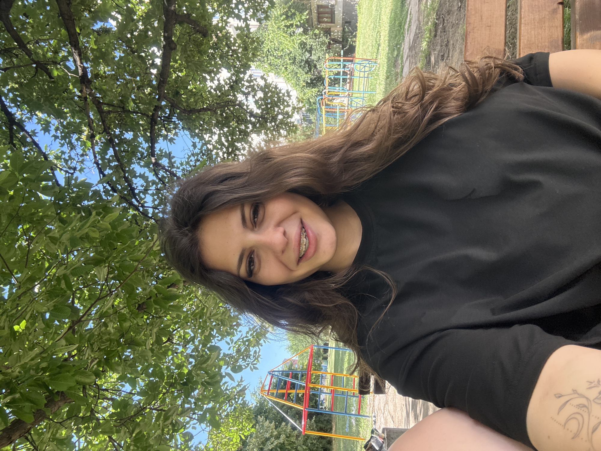 A young woman with long, wavy brown hair and braces is taking a selfie outdoors. She is wearing a black t-shirt and smiling. In the background, there is a colorful playground structure with red, yellow, and blue bars, green grass, trees with lush green leaves, and a building with windows. The scene appears to be set in a park or a residential area with a playground. The sunlight suggests it is daytime. The overall setting could be a neighborhood playground in Plovdiv, Bulgaria.