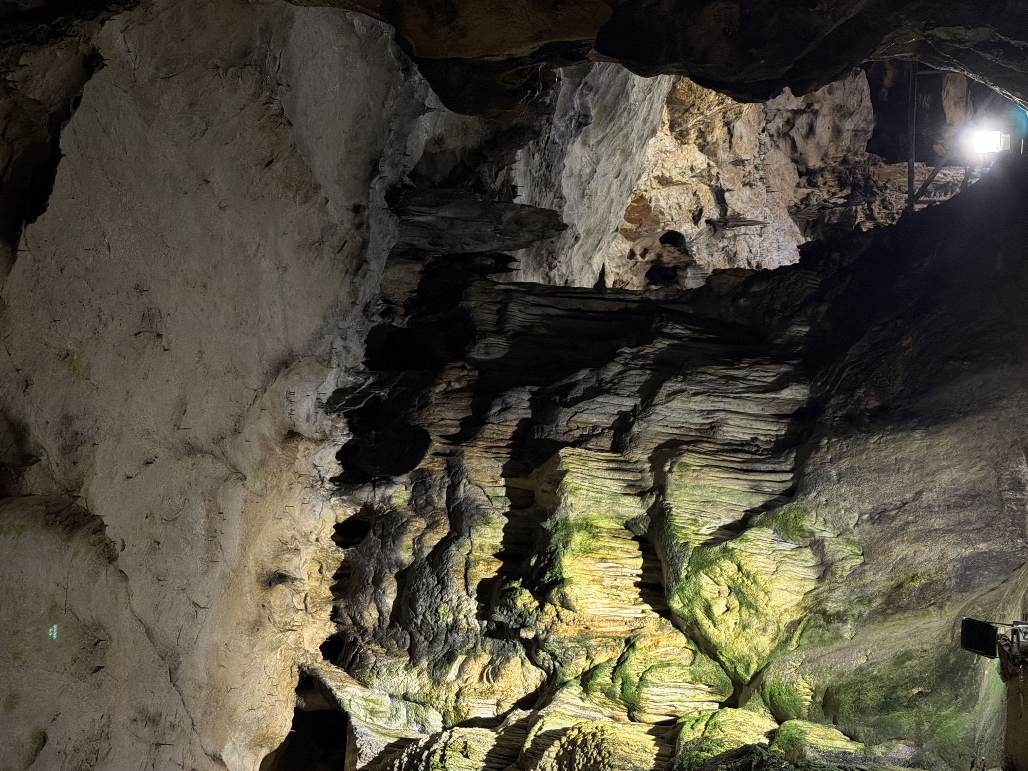 The image depicts the interior of a cave. The cave walls are composed of layered rock formations, some of which are covered in green moss or algae. A light source is visible in the upper right area of the image, illuminating a section of the cave. No people are visible. Location: Unknown, Unknown.