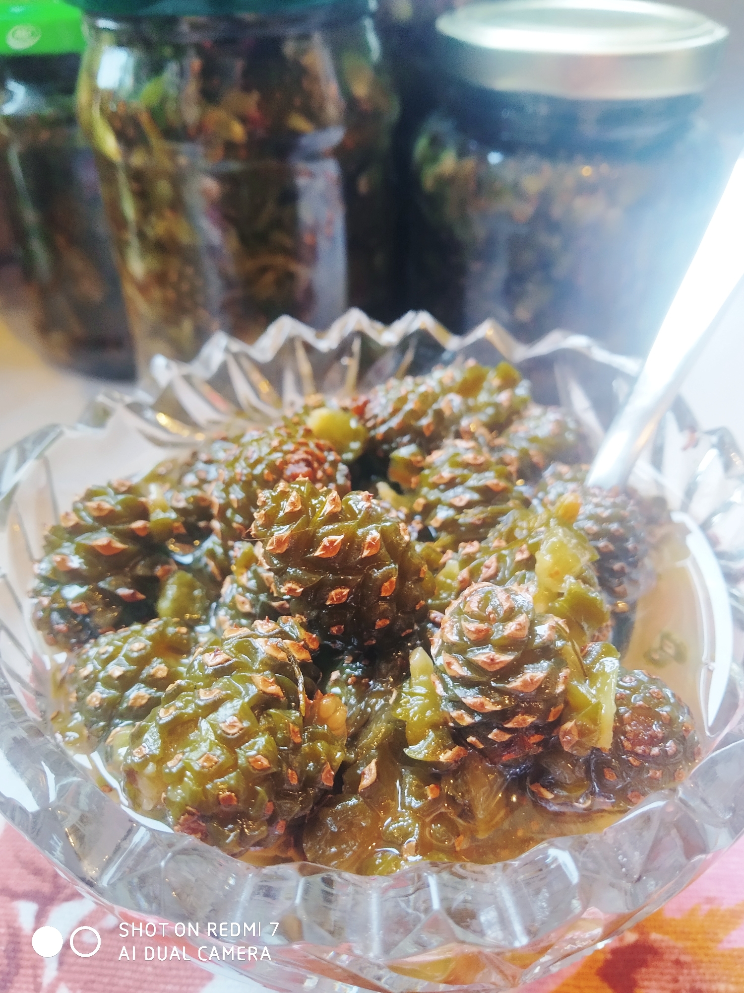 The image shows a glass bowl filled with what appears to be a preparation of young pine cones, likely a jam or preserve, sitting on a patterned surface. A spoon rests in the bowl, suggesting the food is ready to be consumed. In the background, several glass jars are visible, presumably containing more of the same pine cone preparation, which suggests the location is Chaikovsky in Russia. The objects in the background are slightly out of focus, drawing attention to the bowl in the foreground.