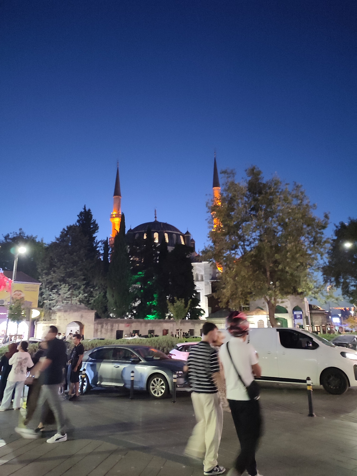The image depicts a street scene in Üsküdar, Turkey, at dusk. In the background, a mosque with two minarets is illuminated, with the minarets lit in orange. A large tree with sparse foliage stands in the mid-ground, obscuring some of the mosque's facade. The sky is a deep blue, transitioning to twilight. In the foreground, several people are walking. To the left, a group of individuals are standing near a building with posters. A silver car is parked on the right side of the street, and a white van is also visible. The street appears to be paved. The overall atmosphere suggests a public area with pedestrian activity during the evening.