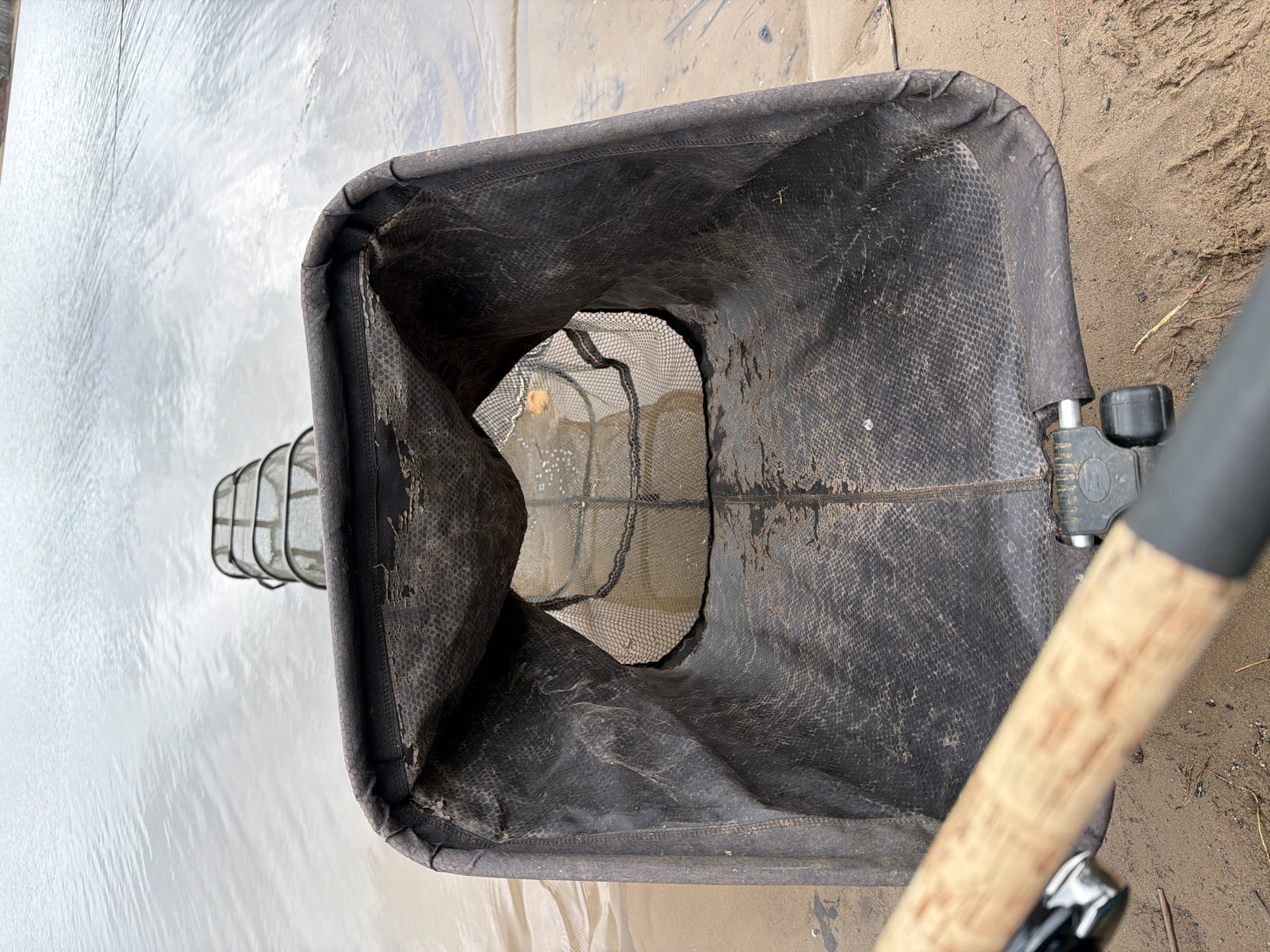 A fishing net is partially submerged in shallow water near a sandy shoreline. The net has a dark, square-shaped frame with a mesh interior, which is partially collapsed or damaged in one area. The handle of the net extends diagonally toward the right side of the frame. The water is a murky light grey and the sand is also greyish. The setting appears to be outdoors, possibly near a river or lake. The image is oriented in a way that looks down into the net. There is no visible evidence of people, interactions, or specific environmental details that would identify it to be in Chaikovsky, Russia.