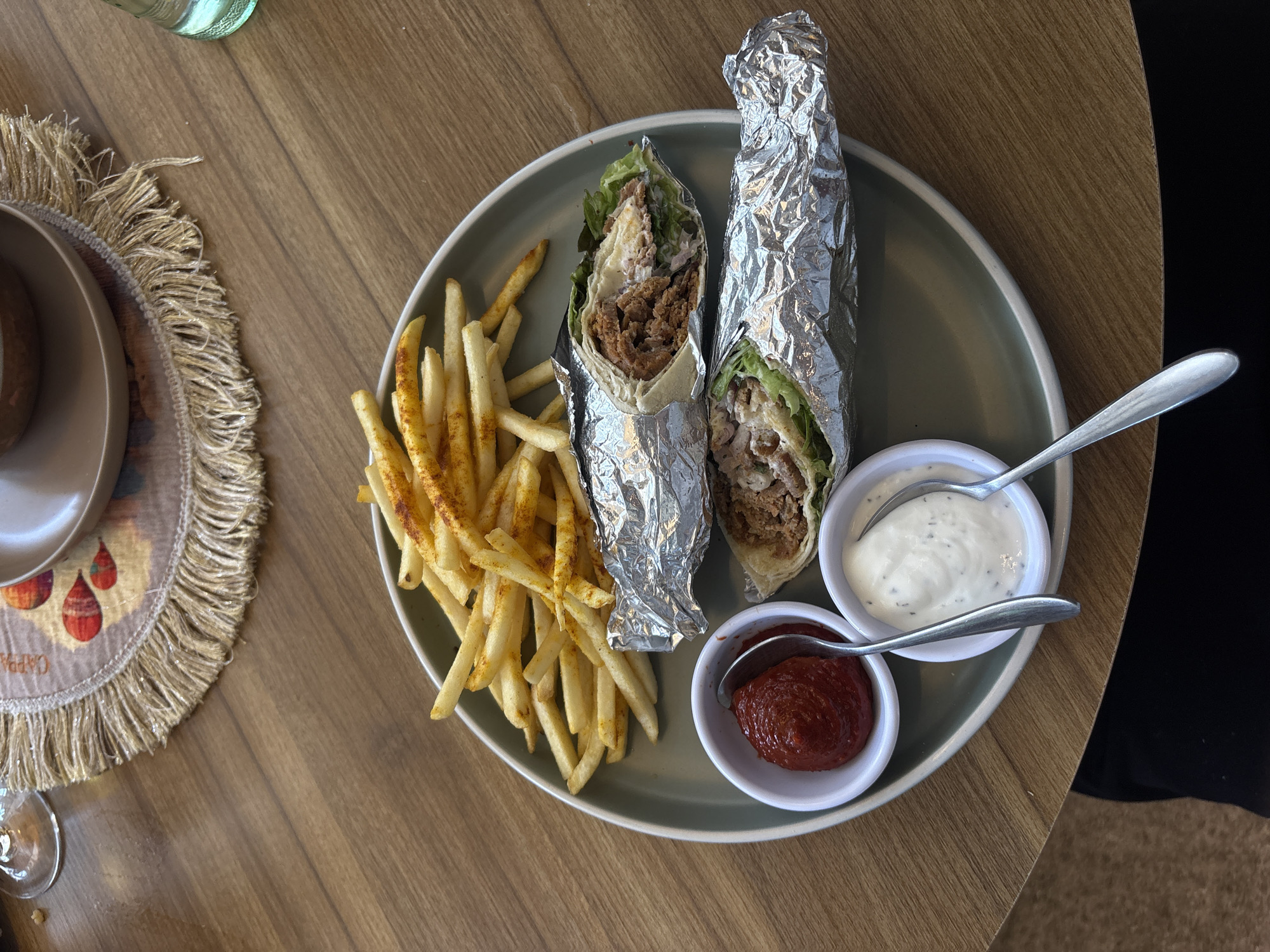 The image displays a close-up, overhead view of a meal served on a wooden table. The central subject is a large, round, light green-grey ceramic plate containing food items. On the plate, a pile of golden-brown french fries seasoned with a reddish-orange spice occupies the upper left quadrant. Below the fries are two elongated wraps, each partially encased in crinkled aluminum foil. The visible portions of the wraps reveal a tortilla filled with lettuce and a substantial amount of finely shredded or minced meat, alongside a white creamy sauce. To the lower left of the wraps, two small, white ceramic ramekins hold dipping sauces: one with a thick red sauce (consistent with ketchup) and another with a white, creamy sauce containing dark specks (likely herbs). A silver spoon rests in each ramekin.
The plate rests on a light brown wooden table featuring a visible grain pattern. In the upper right background, a round placemat with a fringed edge is partially visible. The placemat is light brown with a repeating pattern of reddish-orange, tear-drop shaped motifs and partially discernible text reading "CAPRA". A brown ceramic bowl and its matching saucer are situated on this placemat. A clear, fluted glass, possibly containing water, is also visible on the table surface to the right of the placemat. The lower edge of the image reveals a portion of a person's leg or lap, clad in dark fabric, suggesting a seated individual at the table.
The scene takes place indoors, consistent with a casual dining establishment within Heroica Puebla de Zaragoza, Mexico. However, the precise street address or specific venue name cannot be determined from the visual evidence. The lighting is even, suggesting ambient or overhead illumination.