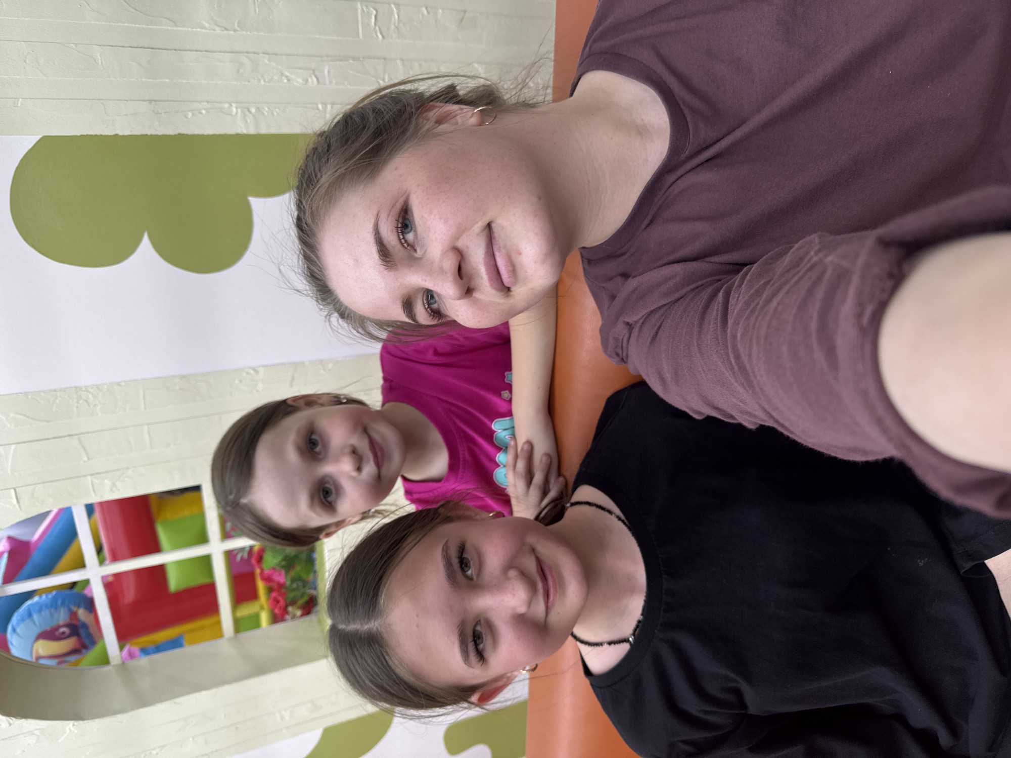 Three young girls are posing for a selfie. The girl on the bottom is wearing a black T-shirt and a beaded necklace, with her hair pulled back. The middle girl is wearing a pink T-shirt, while the girl on top is wearing a maroon long-sleeved shirt. They are indoors, in what appears to be a play area with colorful decorations and an inflatable structure visible through a window in the background. The exact location is Chaikovsky, Russia.