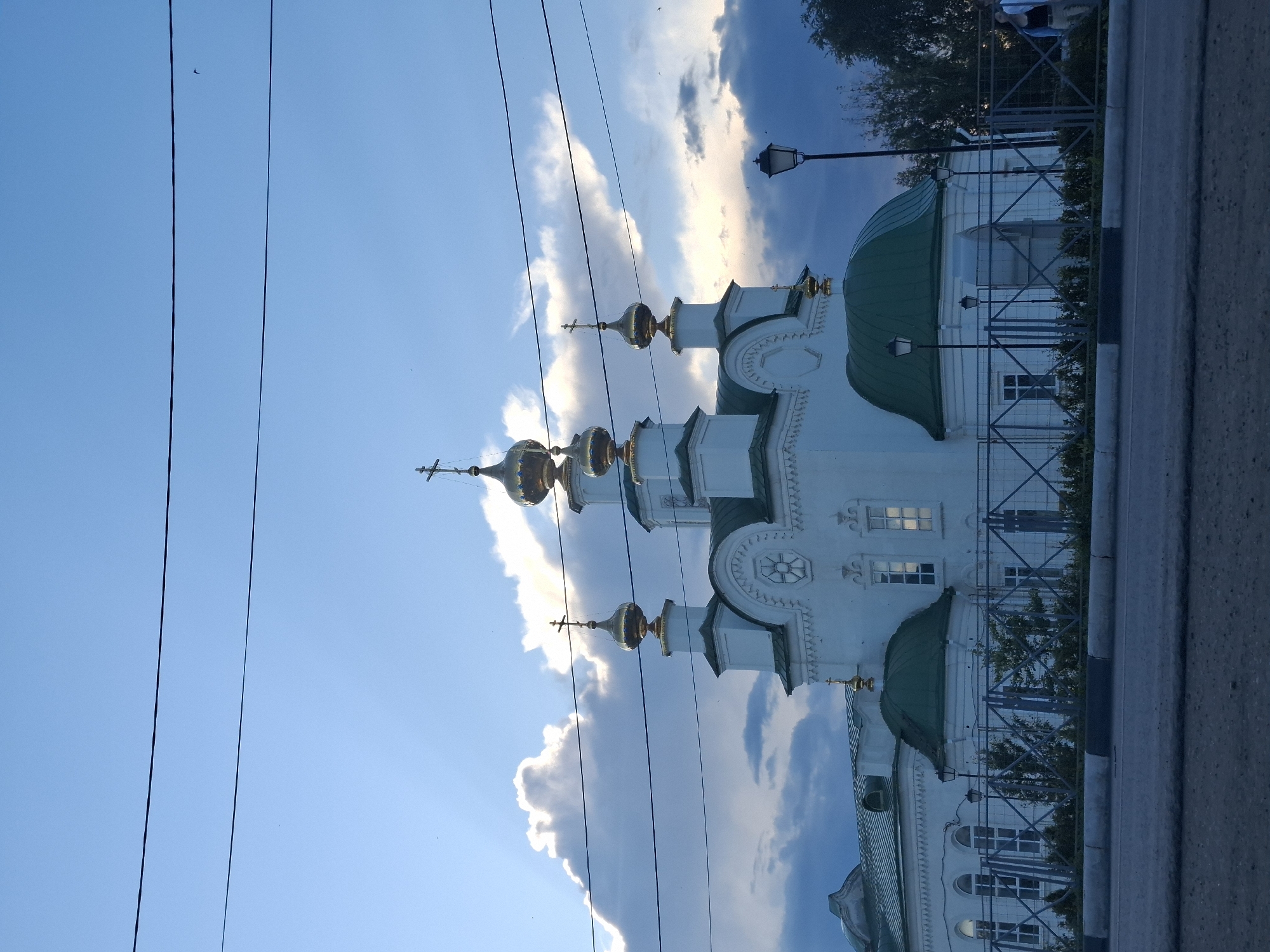 The image captures an Orthodox church in Kungur, Russia, characterized by its white walls and prominent golden domes, topped with crosses. The sky above is a dramatic blend of blue with illuminated white clouds, suggesting either early morning or late afternoon light, with sunbeams piercing through the clouds. Two dark power lines are visible stretched across the upper portion of the frame. The church is enclosed by a metal fence, behind which low bushes and trees are present. A street lamp stands to the right of the church, and a road is in the foreground. A few individuals are partially visible on the far right, near the fence, suggesting a public space. The overall impression is one of a serene and possibly historically significant religious site against a dynamic sky.