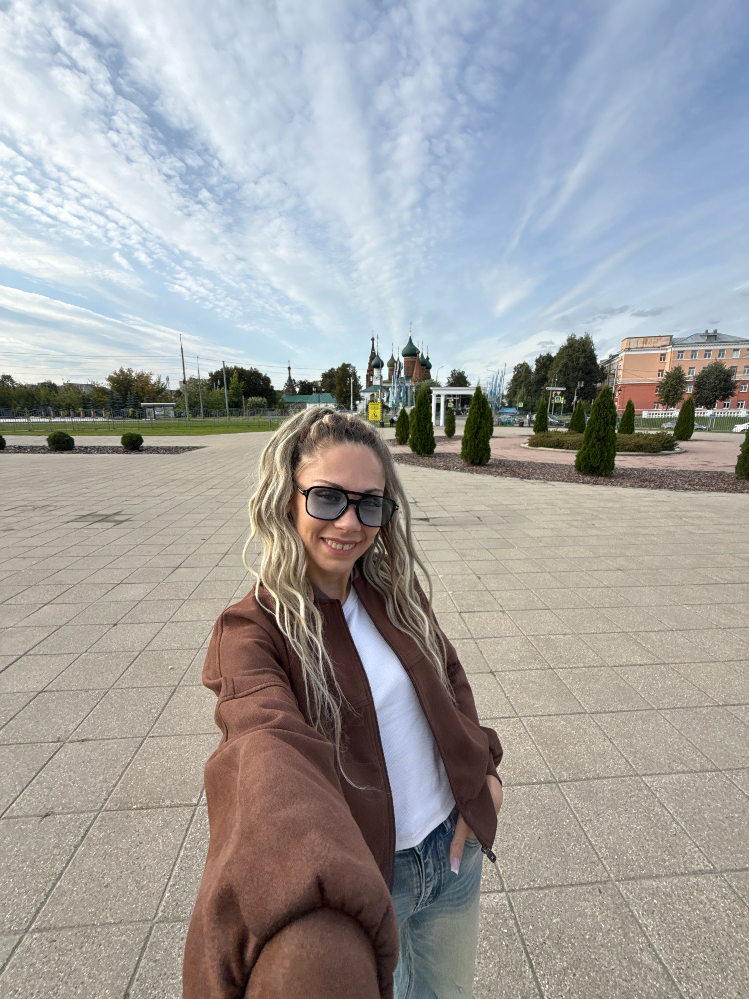 A young woman with blonde hair styled in a high ponytail and wearing large sunglasses, a brown zip-up jacket over a white t-shirt, and light-wash jeans is taking a selfie. She is standing on a paved area with a pattern of square tiles. In the background, there are several trees, some manicured conical evergreen trees, a building with a red and tan facade, and behind the trees, a collection of onion-domed churches with green roofs. The sky is partly cloudy with wispy white clouds against a pale blue background. The overall scene appears to be an outdoor public space in the city of Korobovo, Russia.