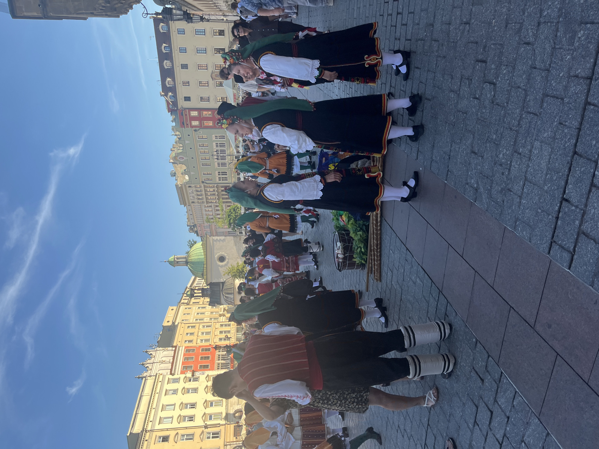 In Krakow, Poland, on the Main Market Square, a group of people in traditional folk costumes are gathered. The square is paved with cobblestones, and colorful historic buildings line the background, under a clear blue sky with wispy clouds. Several women are dressed in black dresses with intricately embroidered hems and bright green headscarves. Some men are also present, one wearing a red vest over a white shirt and black pants with distinctive white leg warmers. The scene suggests a cultural event or performance is taking place. In the foreground, a drum is visible, along with bundles of sticks, hinting at musical accompaniment or props for a dance. The overall atmosphere is lively and festive.