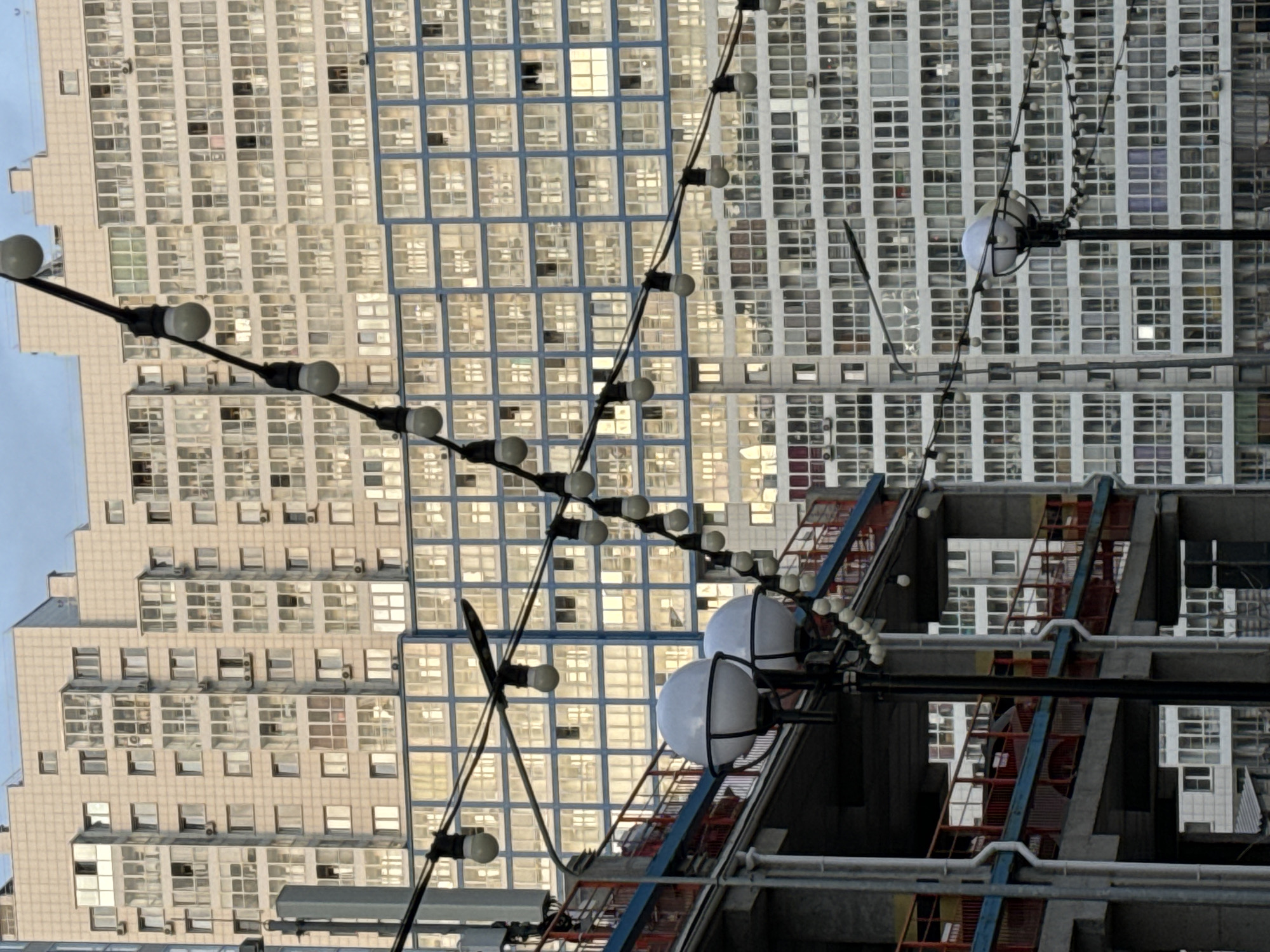 The image depicts a street scene in Krasnoyarsk, Russia, against a backdrop of dense, multi-story buildings. The buildings, which appear to be residential or commercial, have a grid-like facade composed of numerous windows. Hanging across the scene are strings of lights, with the bulbs appearing as spherical white objects against the geometric patterns of the buildings. To the right, a partial view of a building under construction or renovation is visible, featuring a scaffold-like structure with elements in red and blue. The scene is captured in daylight, with the light reflecting off the windows of the buildings. The overall environment conveys a sense of urban density and ongoing construction or maintenance activity.