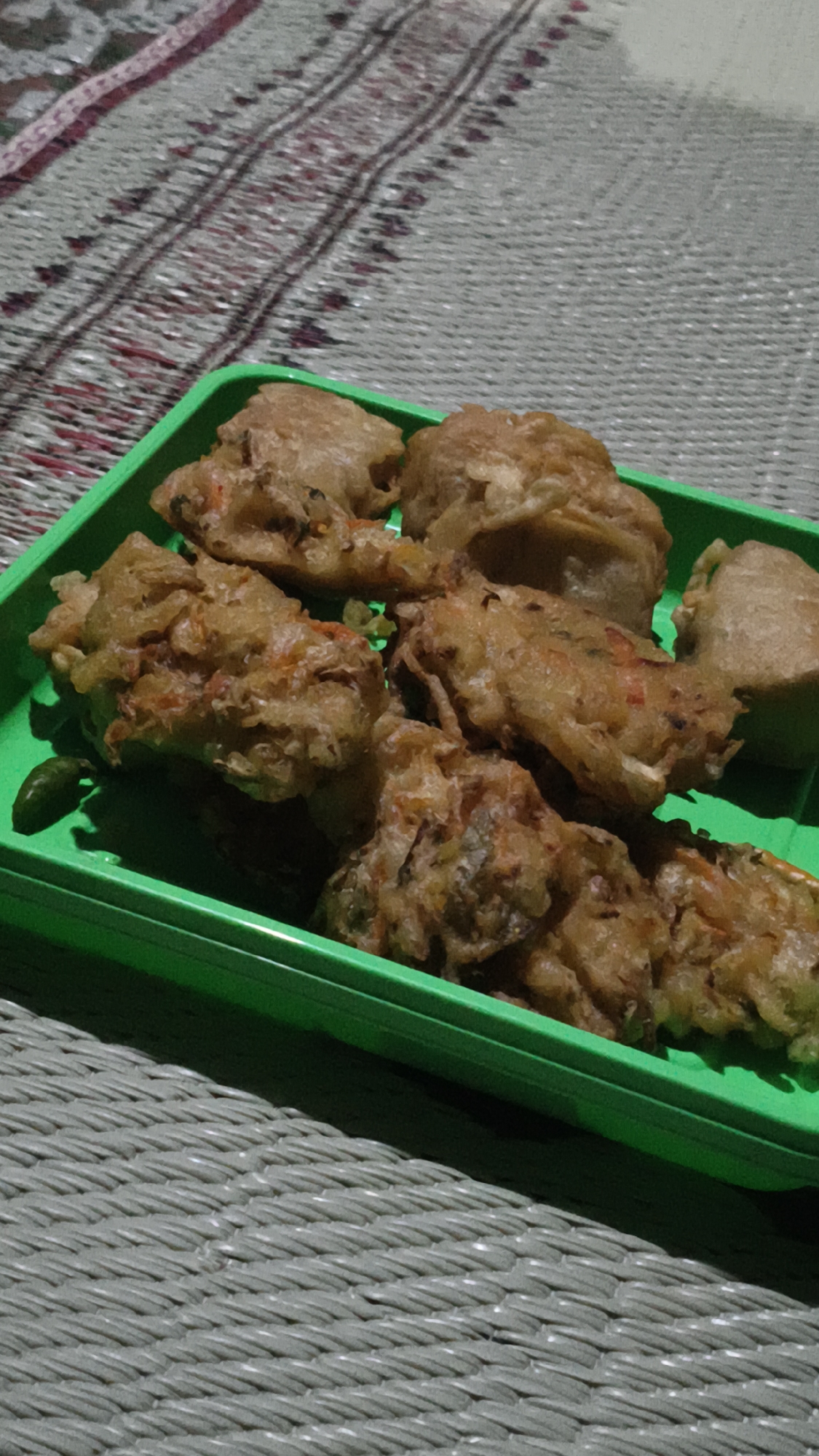 A close-up, overhead shot displays a bright green plastic tray filled with several pieces of fried food, likely a savory fritter or pastry, possibly bakwan, common in Indonesia. The food is golden brown and appears to contain vegetables like shredded carrots and possibly leafy greens. A single, small, green chili pepper is visible to the left of the tray. The tray rests on a woven mat with a textured surface, possibly made of plastic or natural fibers, featuring a repeating linear pattern. In the background, another woven surface with a more intricate, colorful striped pattern is partially visible. The lighting is dim, creating shadows that suggest an indoor setting or evening light. The overall scene evokes a sense of a home-cooked meal or snack.