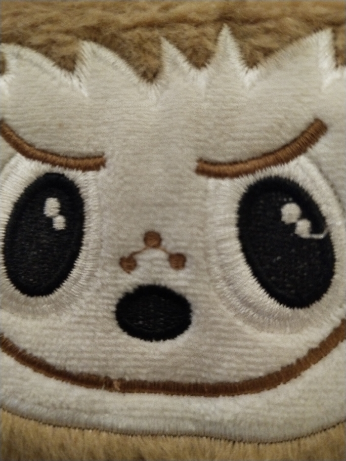 A plush toy's face fills the frame. The toy has white fur with light brown trim. Large black eyes with white reflections sit beneath embroidered brown eyebrows. A small "Y" shaped nose is centered above a black oval mouth. The toy's face has a slightly disgruntled or unimpressed expression, accentuated by the downturned mouth and the brow shape. Brown fur borders the top of the face and the bottom.