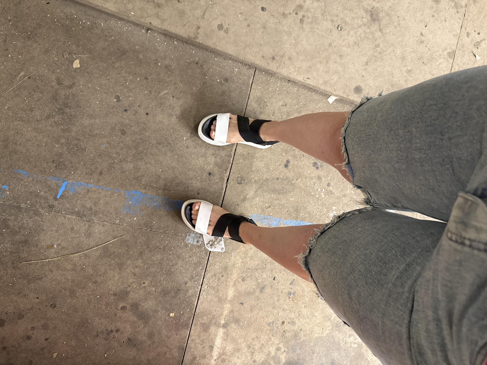 The image shows a pair of feet wearing black and white sandals and denim shorts. The person is standing on a concrete surface with blue paint marks. The scene appears to be outdoors, possibly a sidewalk or an urban area. The lighting suggests it could be daytime, with natural light casting shadows. There are no people fully visible, only the lower part of legs and feet. The focus is on the footwear and clothing, which appear casual. The concrete shows signs of wear and tear, with some debris and cracks. The overall impression is a candid shot of someone's outfit in an everyday setting.