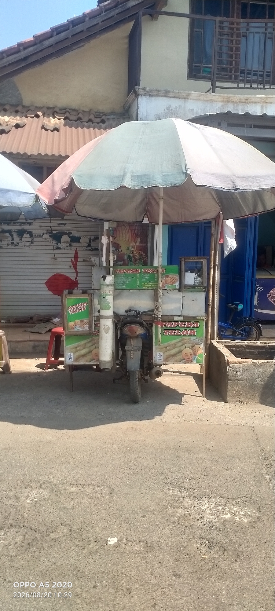 A street vendor cart, possibly selling food, is parked on a paved road in Bugel, Indonesia. The cart is covered by a large, colorful umbrella and features various signs advertising its products. One prominent sign displays "PAPEDA TELOR" with images of food items and cartoon characters. The cart appears to be attached to a motorcycle, suggesting it is mobile. In the background, a building with a tiled roof and shutters is visible. A red plastic stool is placed next to the cart, and a blue bicycle is parked to the right. The scene is illuminated by natural sunlight, casting shadows on the ground.