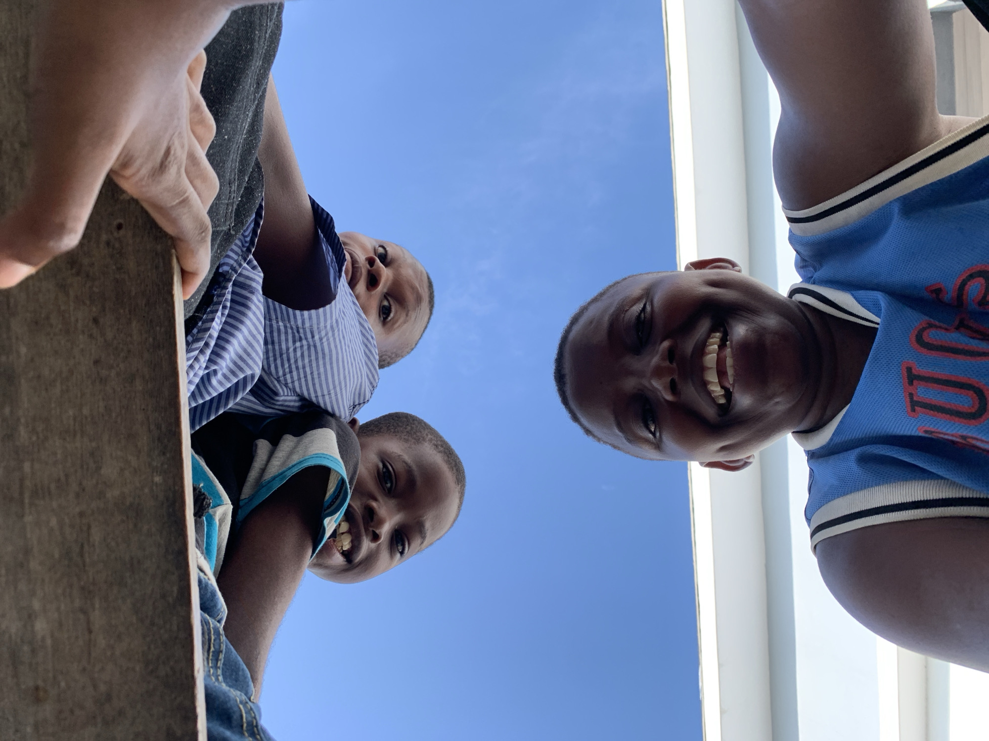Captured from a low-angle perspective, this image shows three young boys looking down towards the camera against a clear, bright blue sky in Ibasa, Nigeria. On the right side, one boy, wearing a blue and white basketball jersey with an illegible red text, smiles broadly, revealing his teeth. On the left, a rustic wooden surface occupies the foreground, with visible hands gripping it. Behind this surface, two other boys are visible. The boy positioned higher wears a blue and white striped button-up shirt and has a neutral, curious expression. Below him, the third boy, in a striped t-shirt (blue, black, and grey) and denim, also smiles. The background is an expanse of cloudless blue sky, indicating clear, sunny daytime weather. A narrow white vertical structure is visible on the far right. The collective gaze and smiling faces suggest a joyful and engaging interaction.
